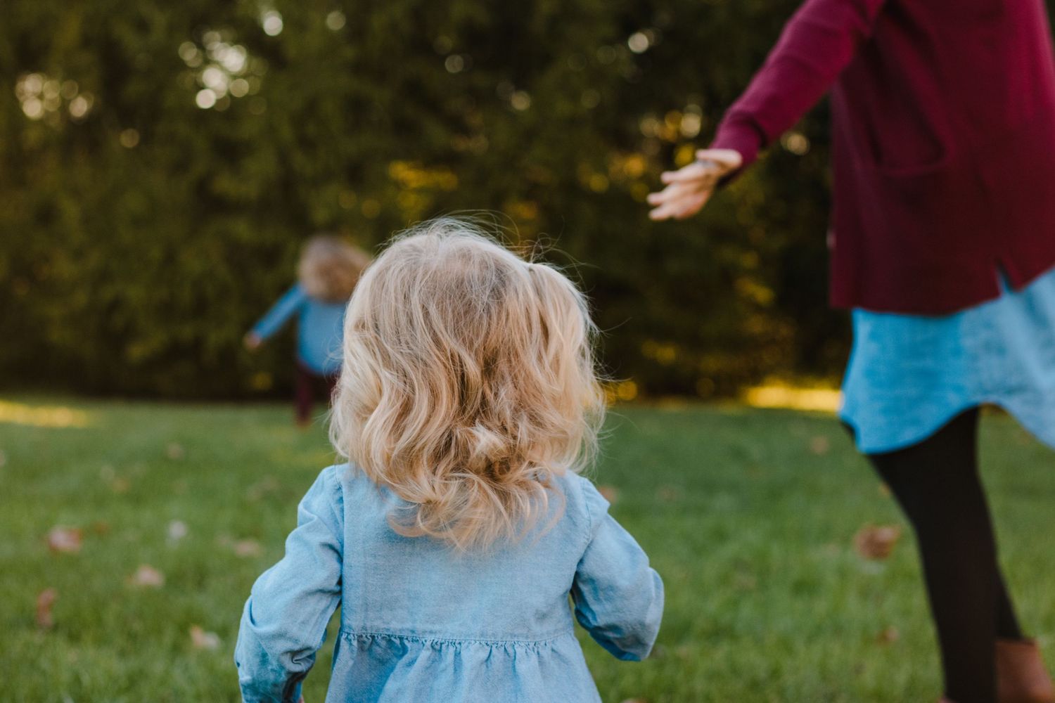 Young blonde toddler girl in denim dress running toward woman in park.