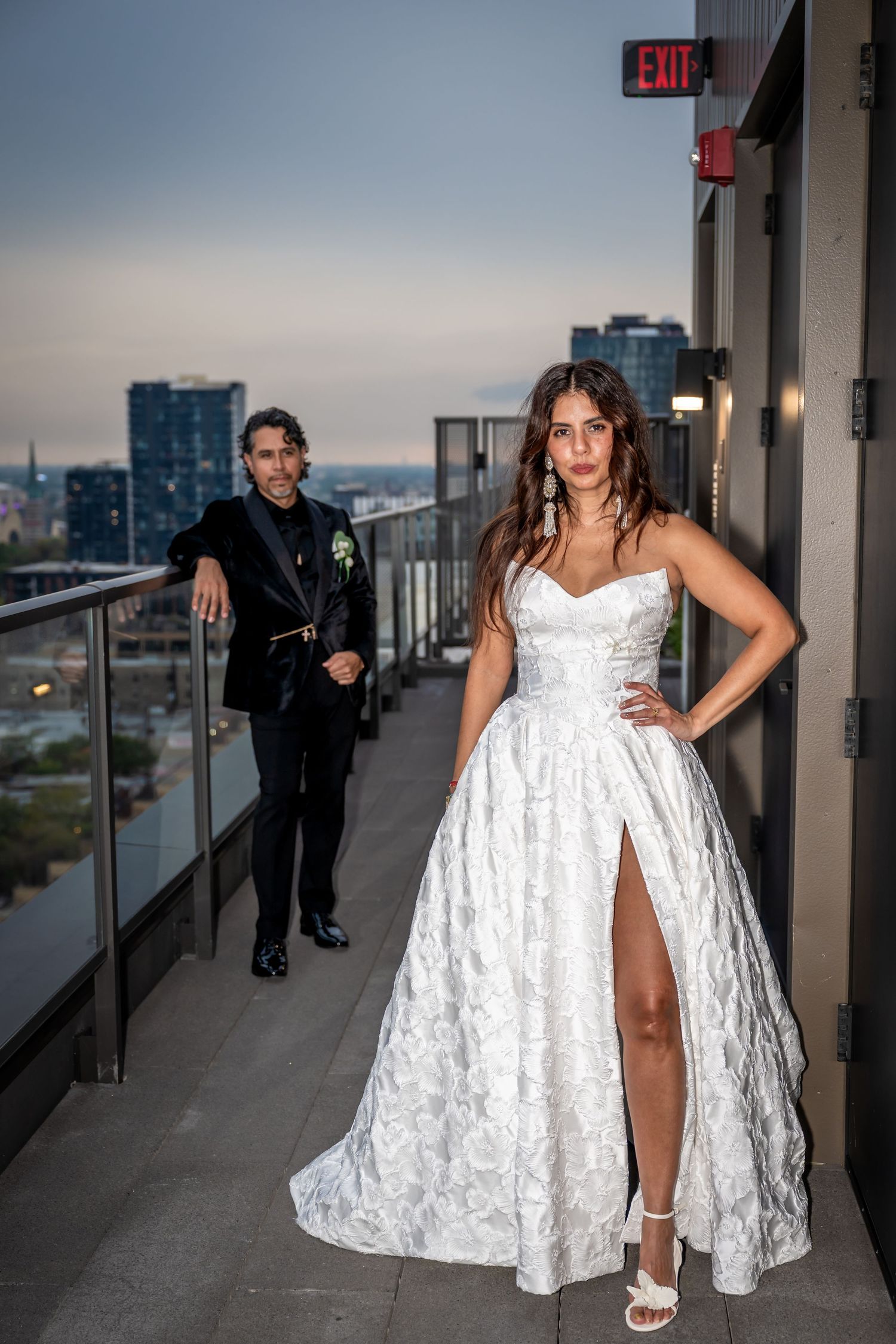 Bride in a white strapless ball gown with slit poses on a rooftop balcony at dusk with groom in black suit behind her.