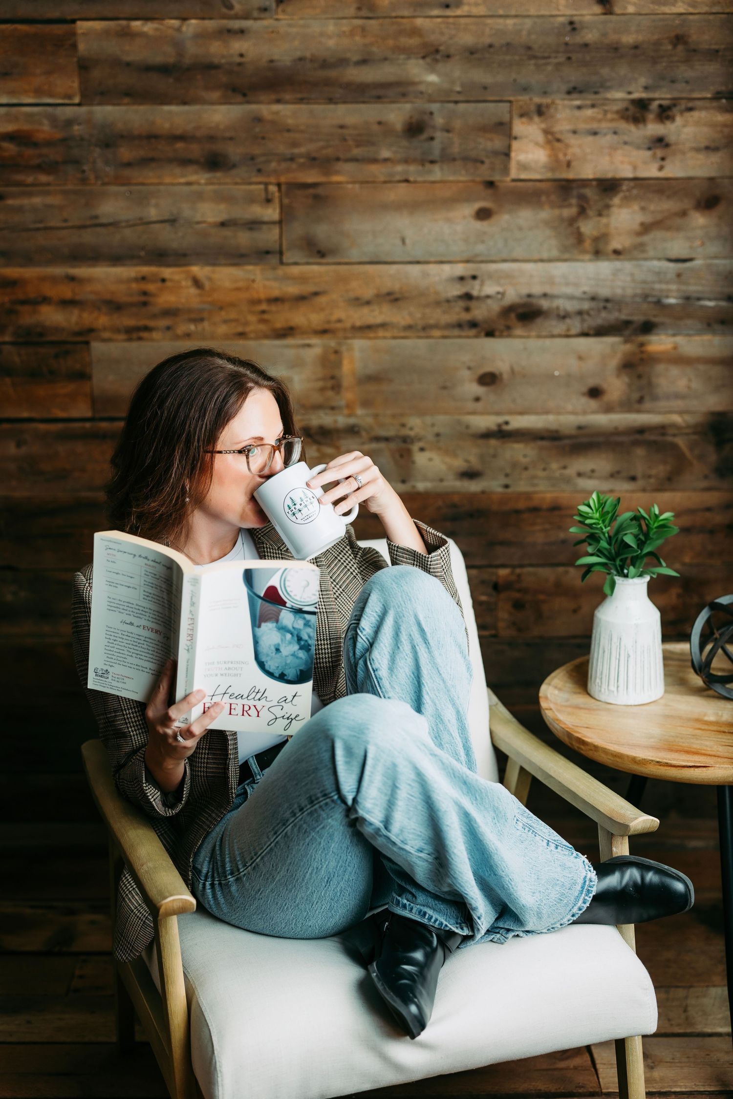 Cozy reading scene with person wrapped in blue blanket holding coffee and book against rustic wooden wall background.