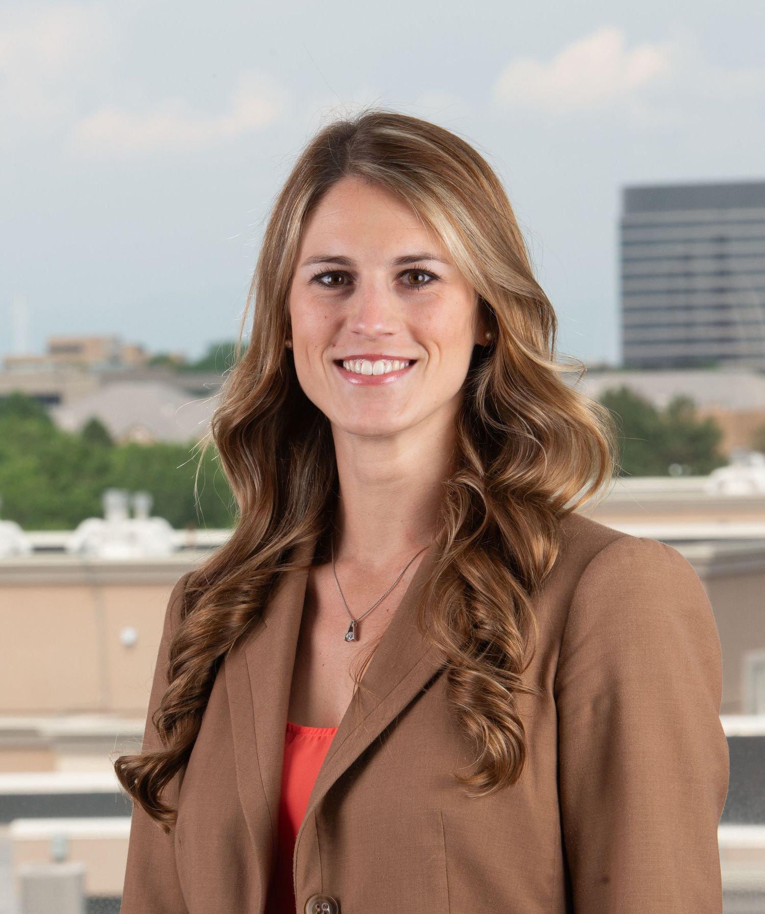 Denver Professional portrait of someone in a brown blazer and coral top against a city backdrop.