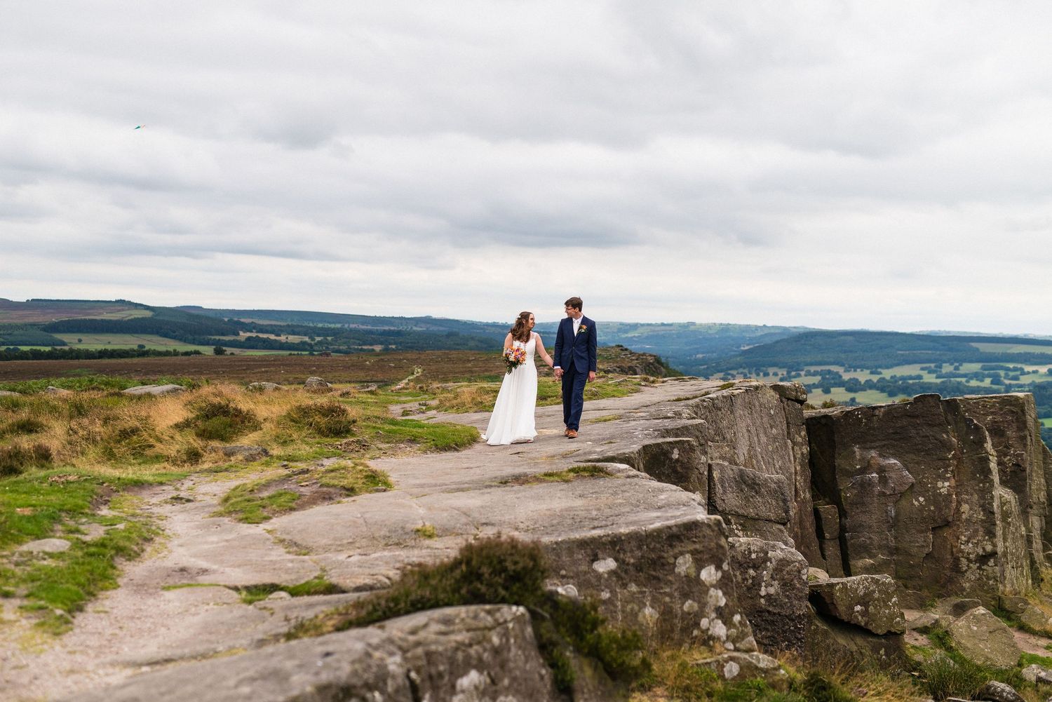 A couple walks along a scenic rocky cliff edge with rolling hills and countryside in the background on an overcast day.