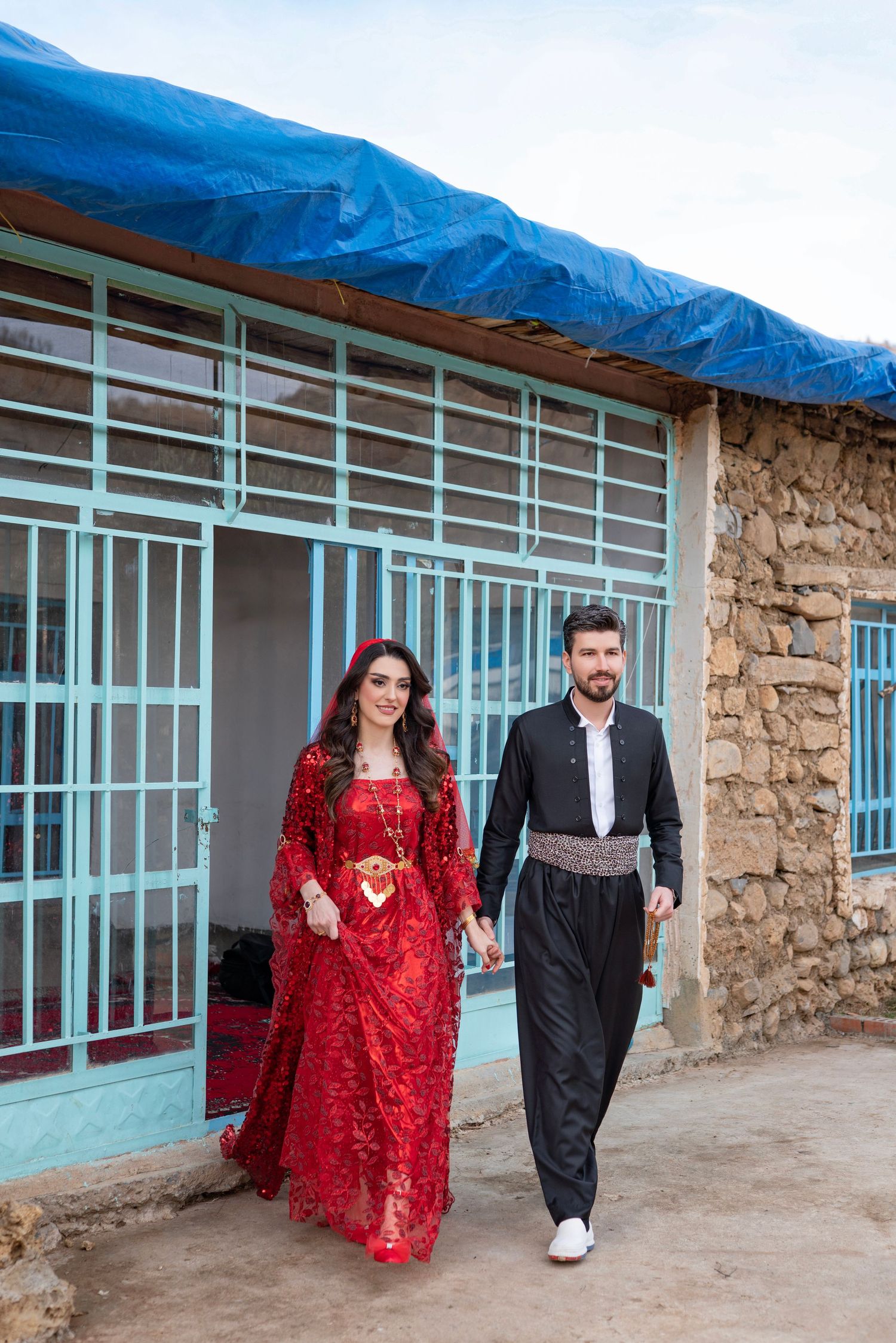 A couple walks together outside a rustic building with turquoise trim and stone walls under a bright blue tarp covering.