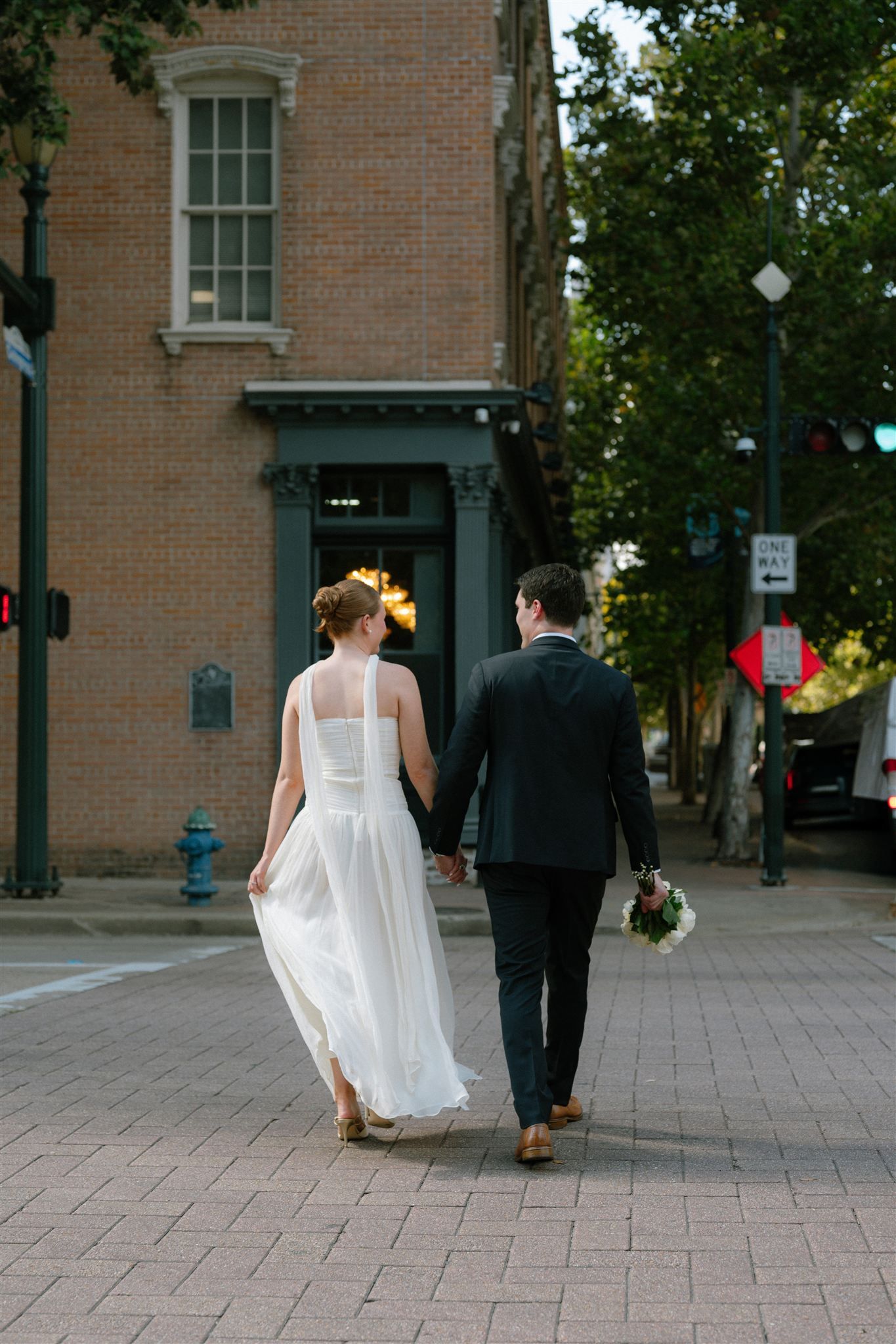 A couple in wedding attire walking together on a brick sidewalk in an urban setting with historic buildings.