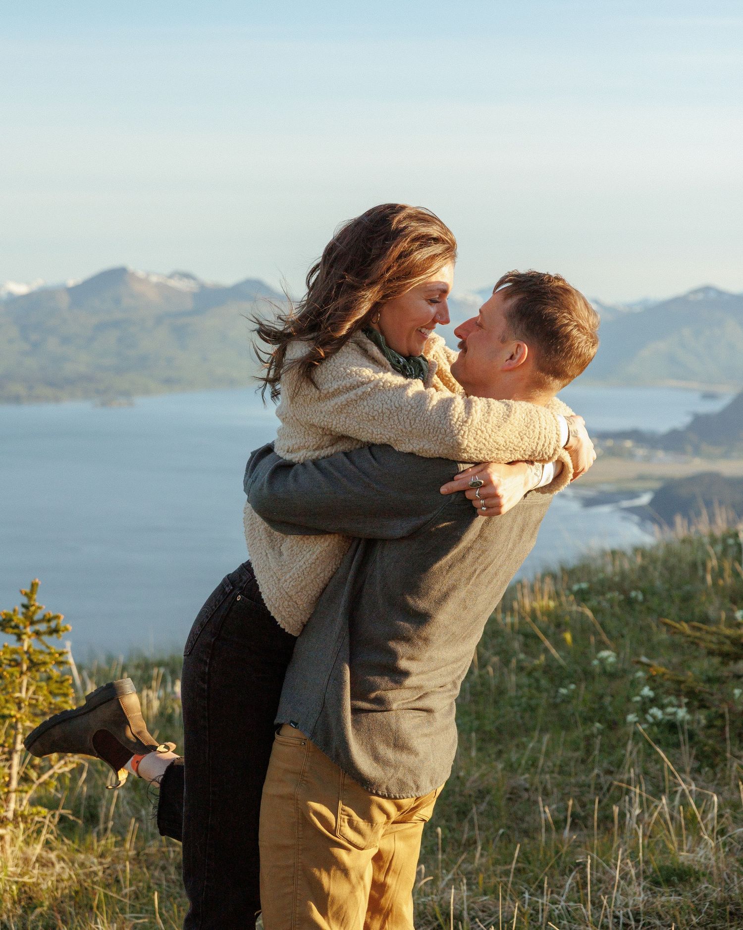Two silhouettes stand together on a mountain ridge at sunset with evergreen trees and wildflowers in the foreground.