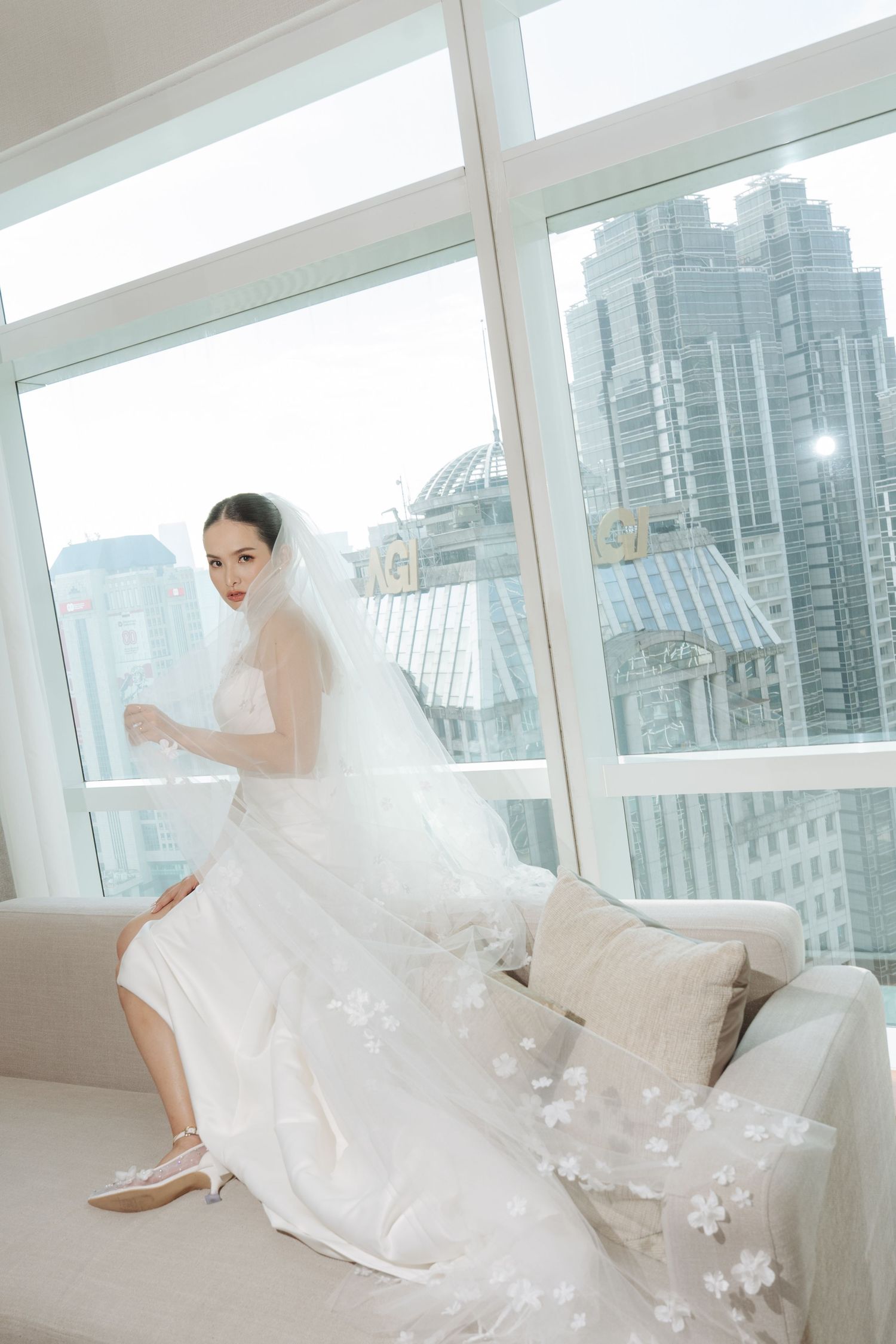 Elegant bride in white gown and floral veil seated on sofa with floor-to-ceiling city skyline windows behind her.