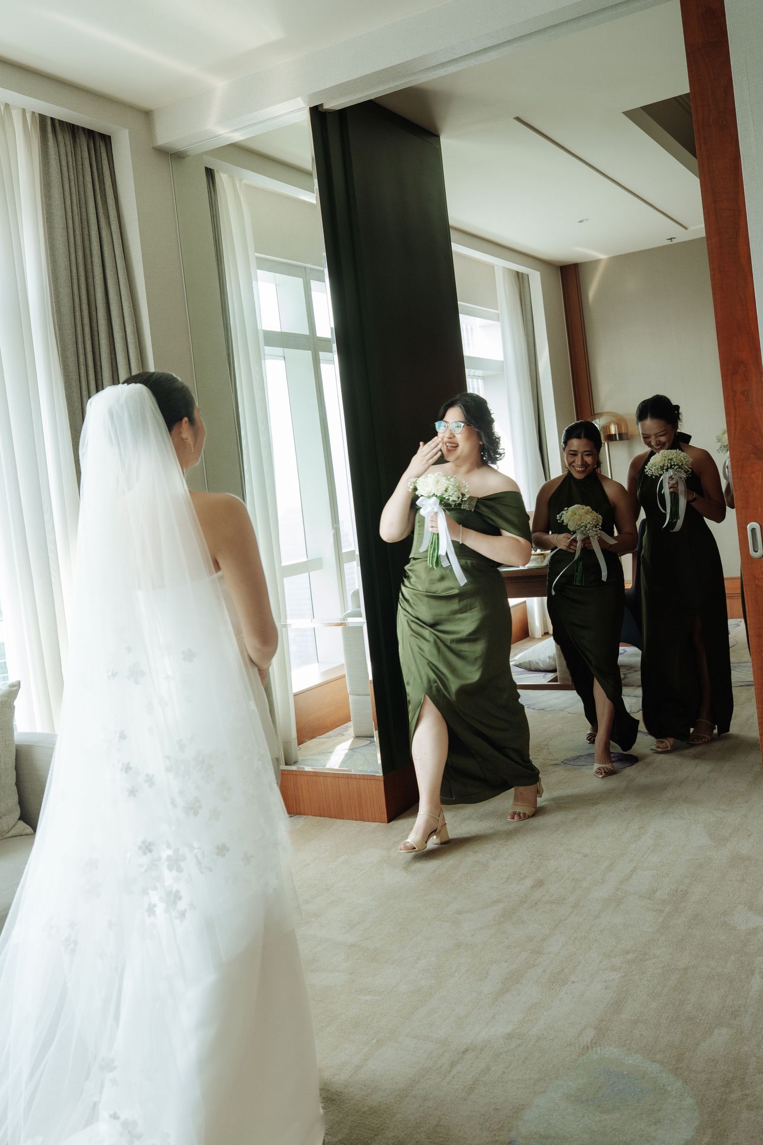 Bridesmaids in olive green dresses react emotionally seeing bride in white lace gown and veil for first time.