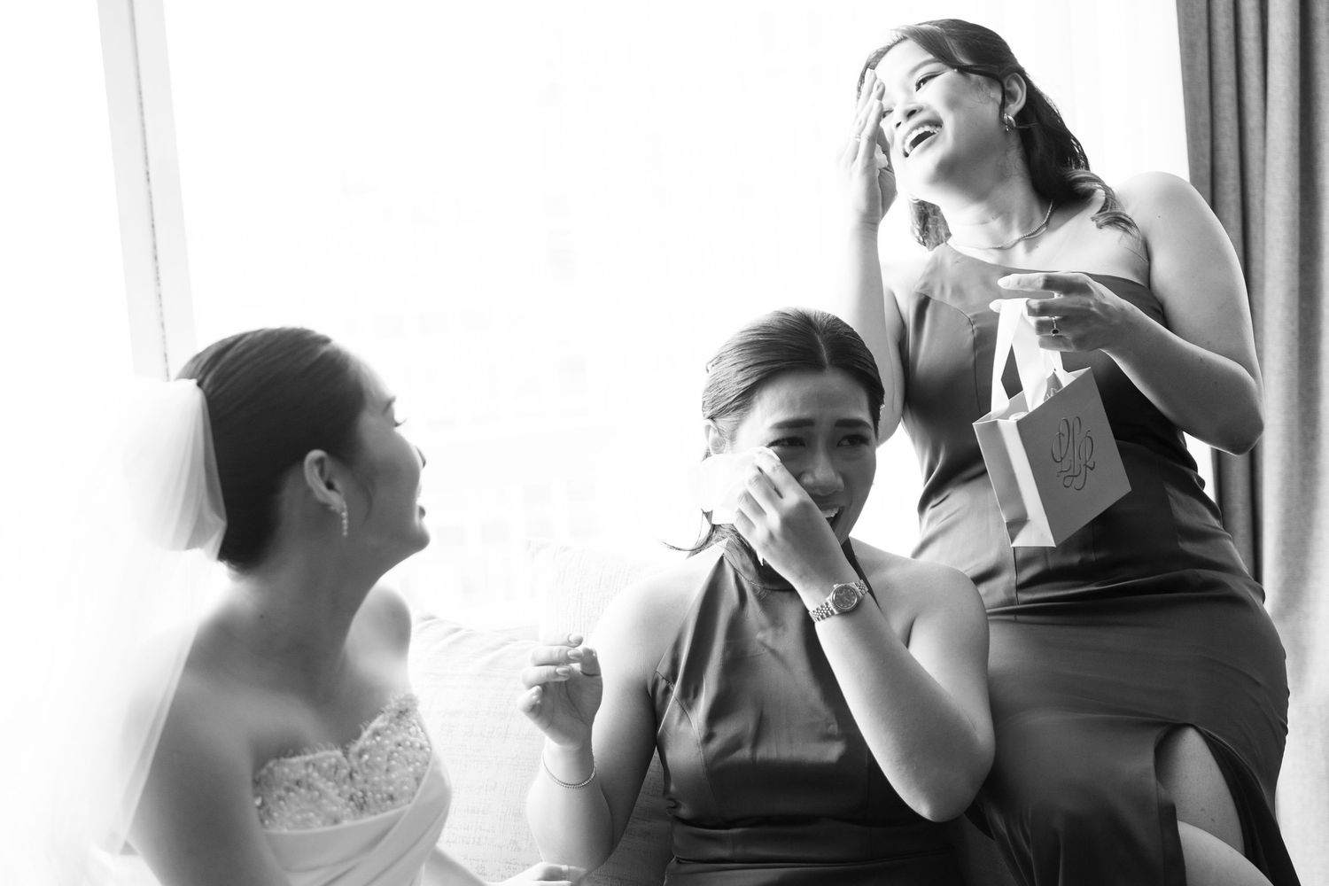 Black and white photo of bride and bridesmaids sharing an emotional moment by a bright window.
