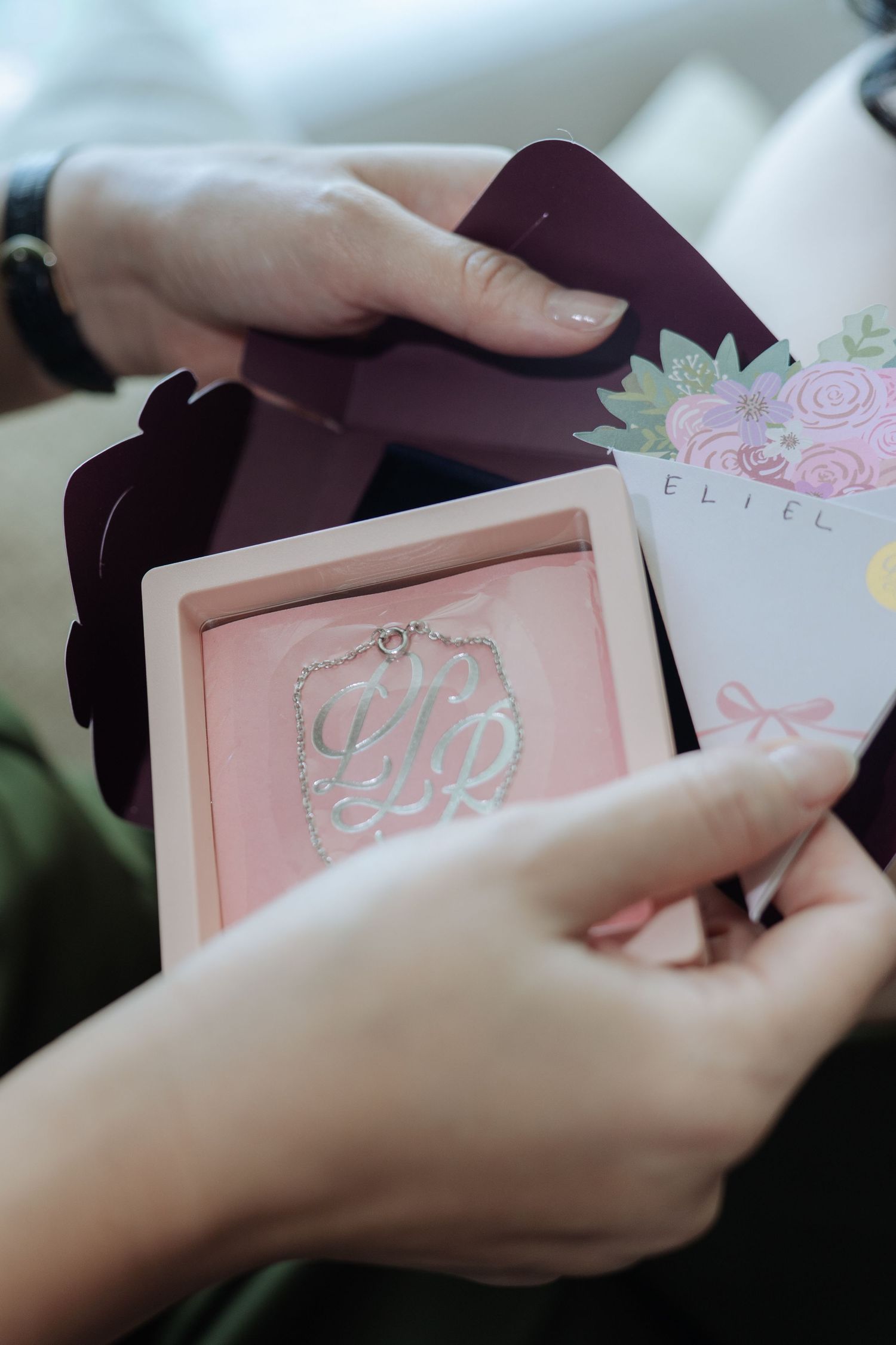 Close-up of hands holding an open gift box with a silver monogram necklace and floral card.