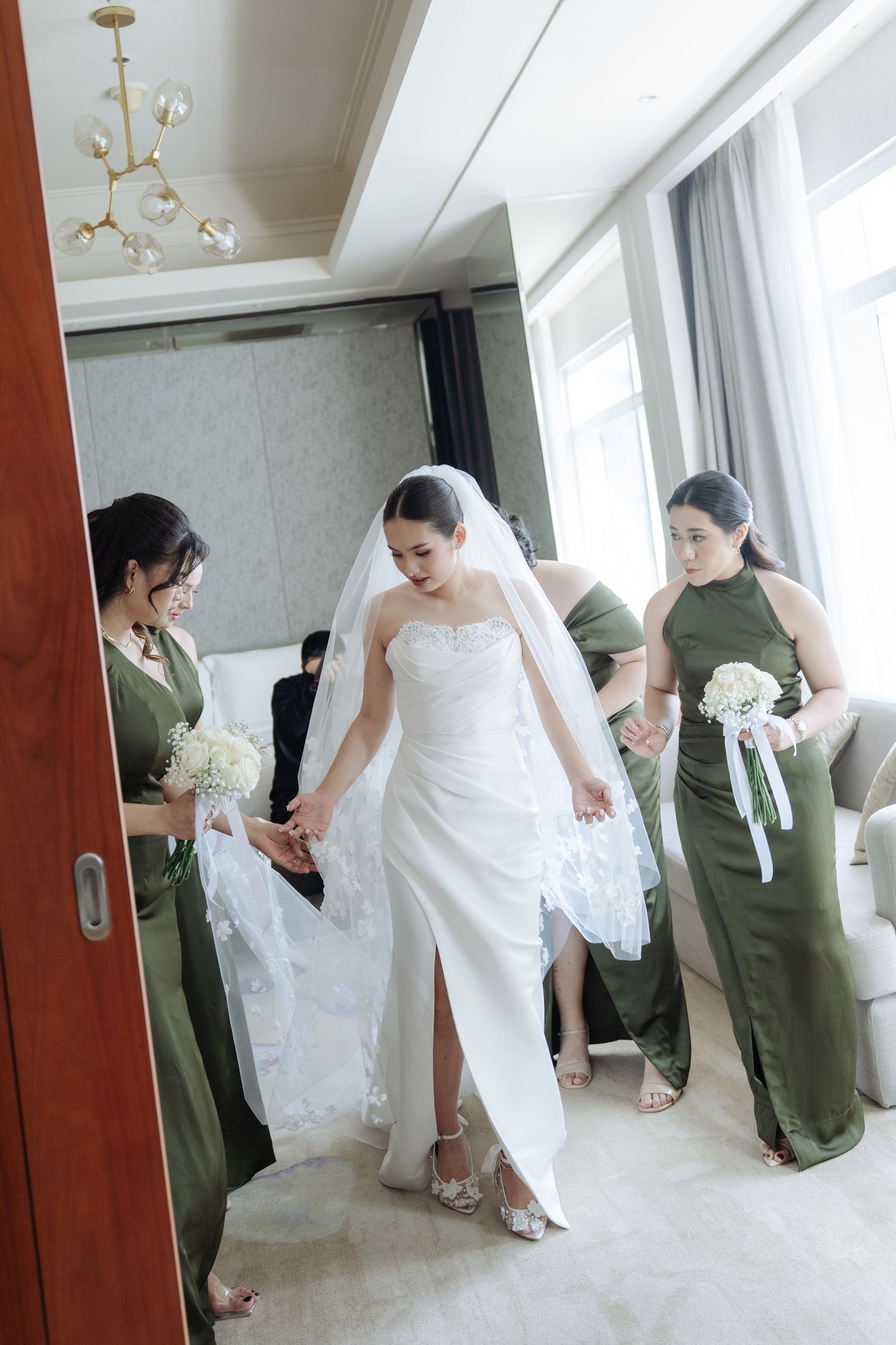 Bride in white gown assisted by bridesmaids in olive green dresses in an elegant hotel room.
