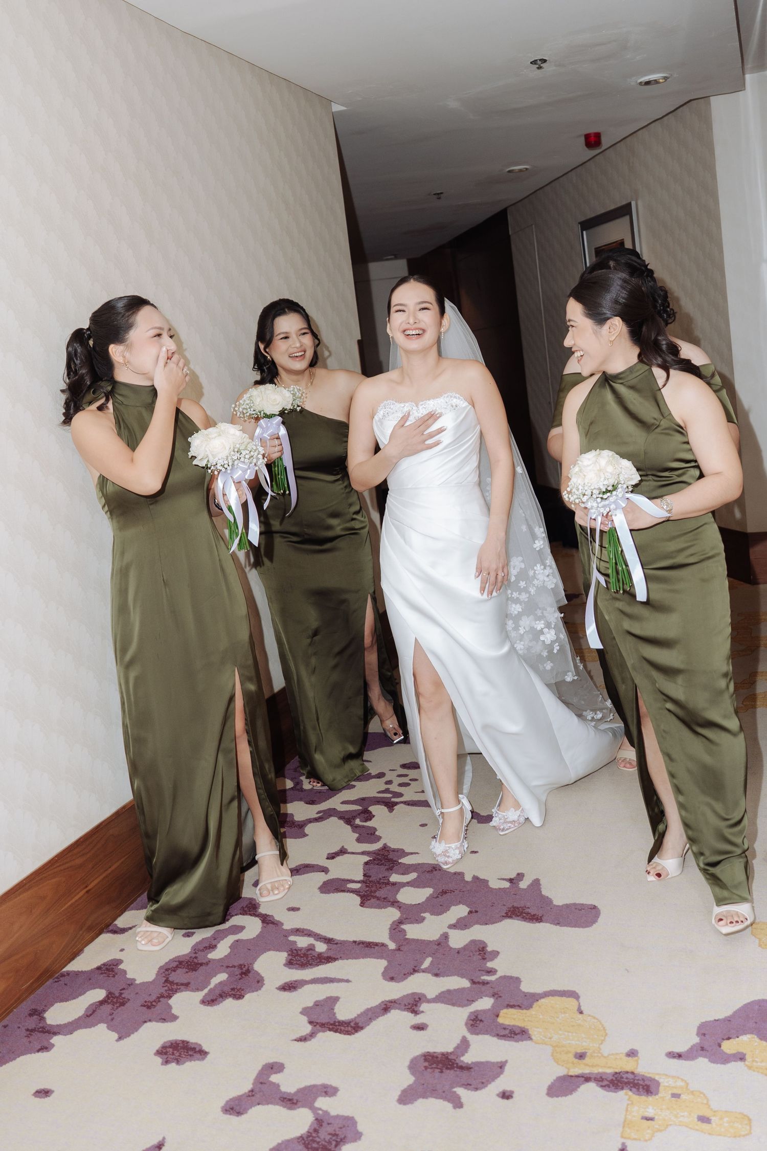 Joyful bride and bridesmaids in olive green gowns walking through a hotel corridor.