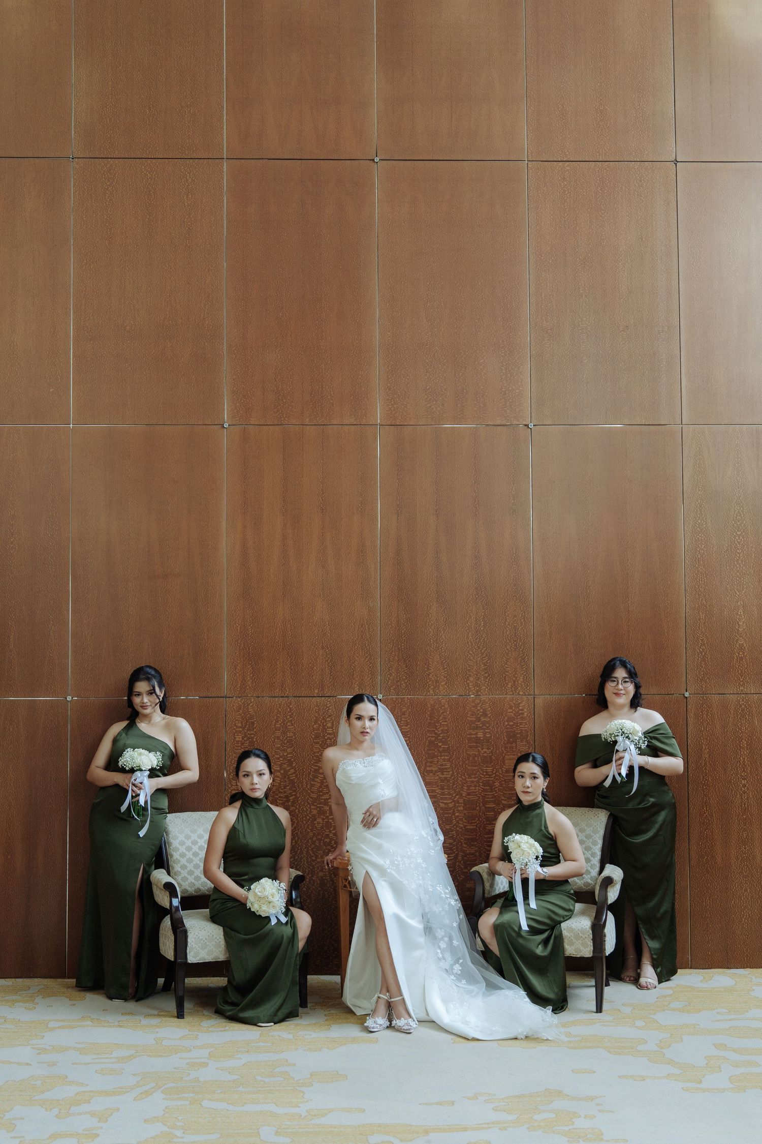 Bride in lace wedding gown poses with four bridesmaids in olive green dresses against a wood wall.