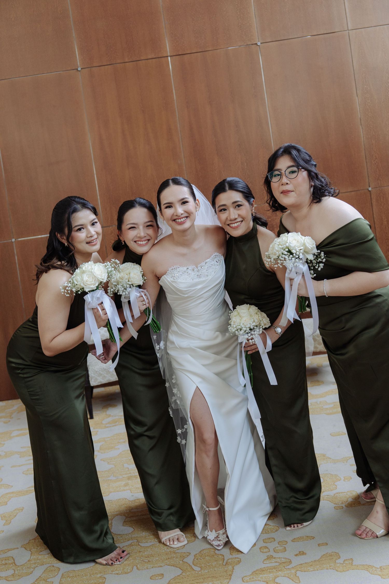 Smiling bride and four bridesmaids in olive satin dresses holding white bouquets pose together.