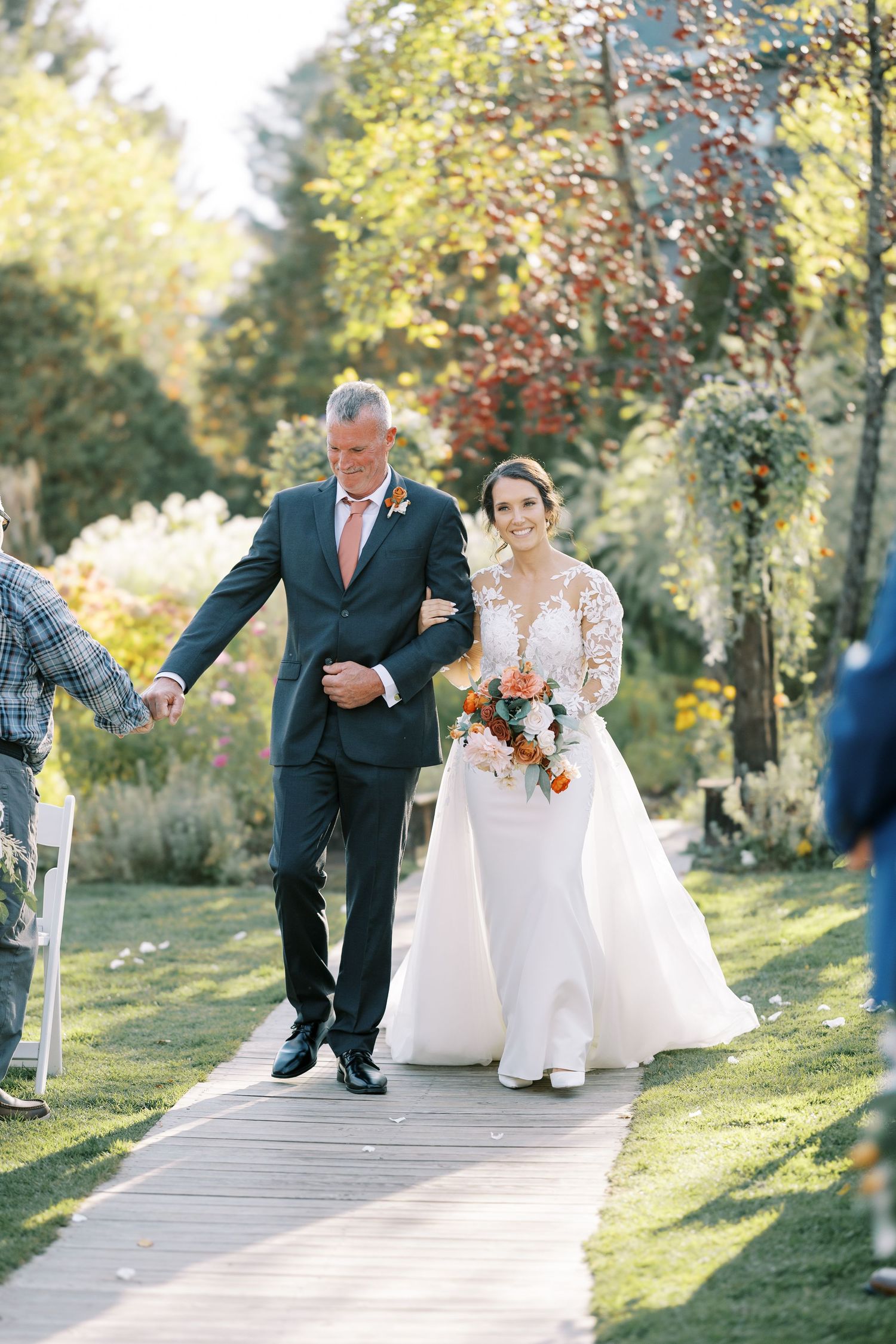A bride and her escort walk down a wooden boardwalk pathway surrounded by autumn foliage on a sunny day.