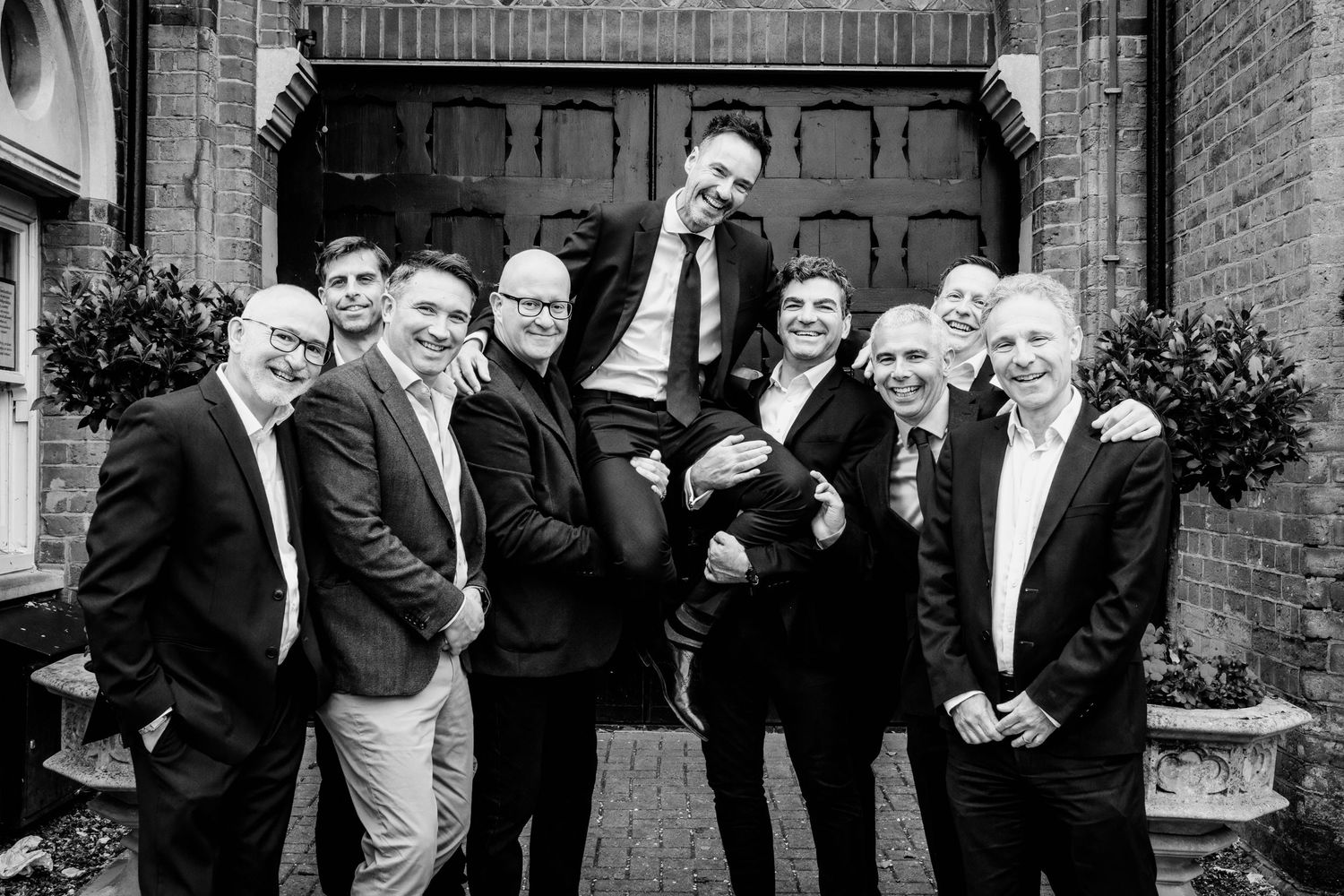 Wedding party poses playfully in black and white photo outside brick building.