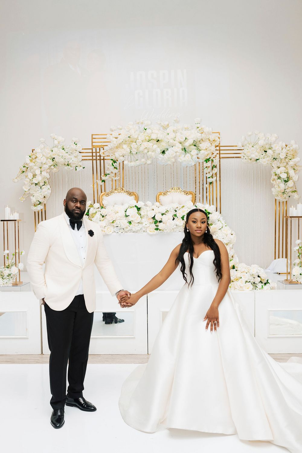 Wedding couple posing against white floral backdrop with gold geometric details.