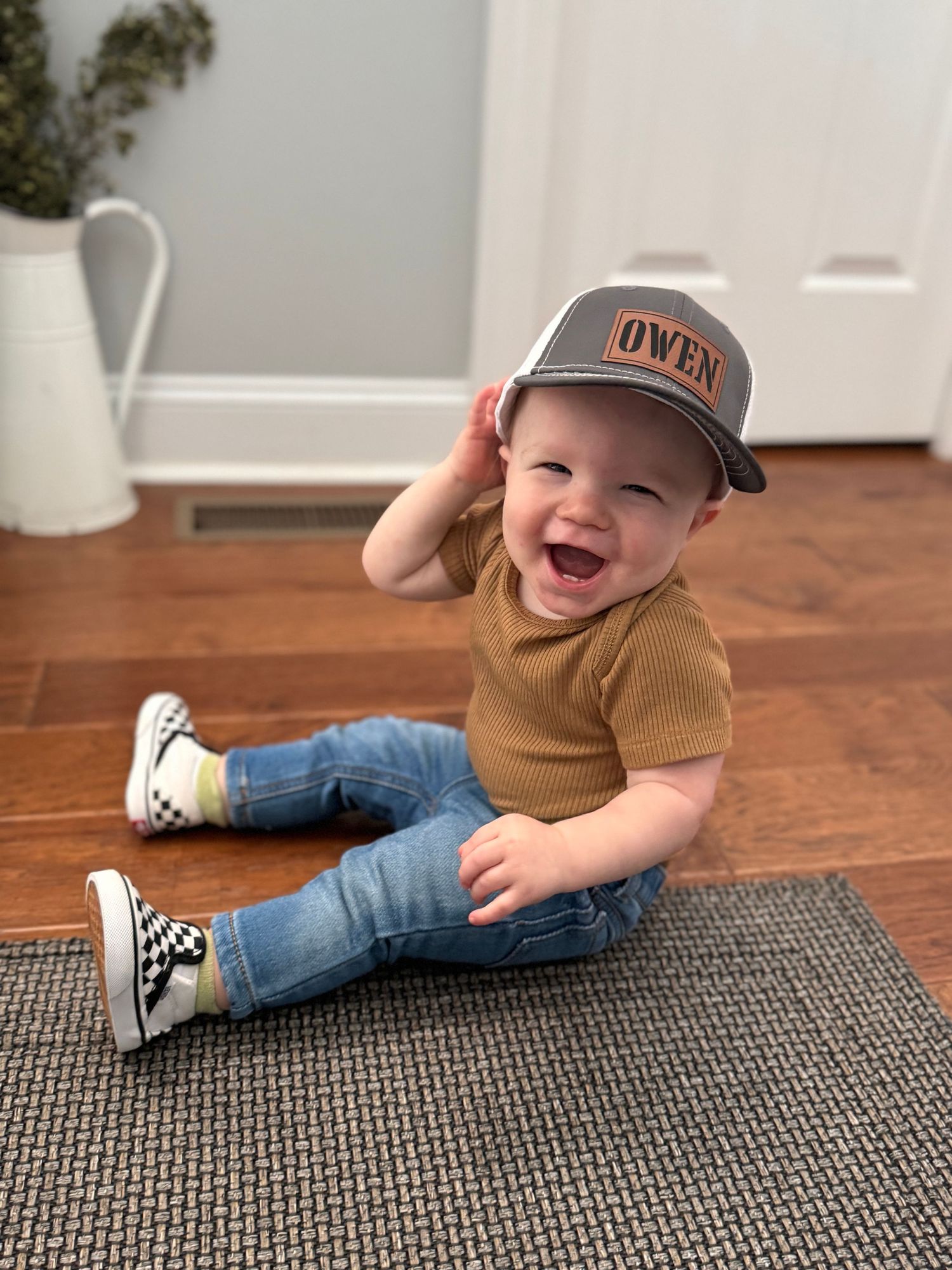 Small child sitting on gray rug wearing baseball cap, brown shirt and jeans with sneakers, expressing joy.