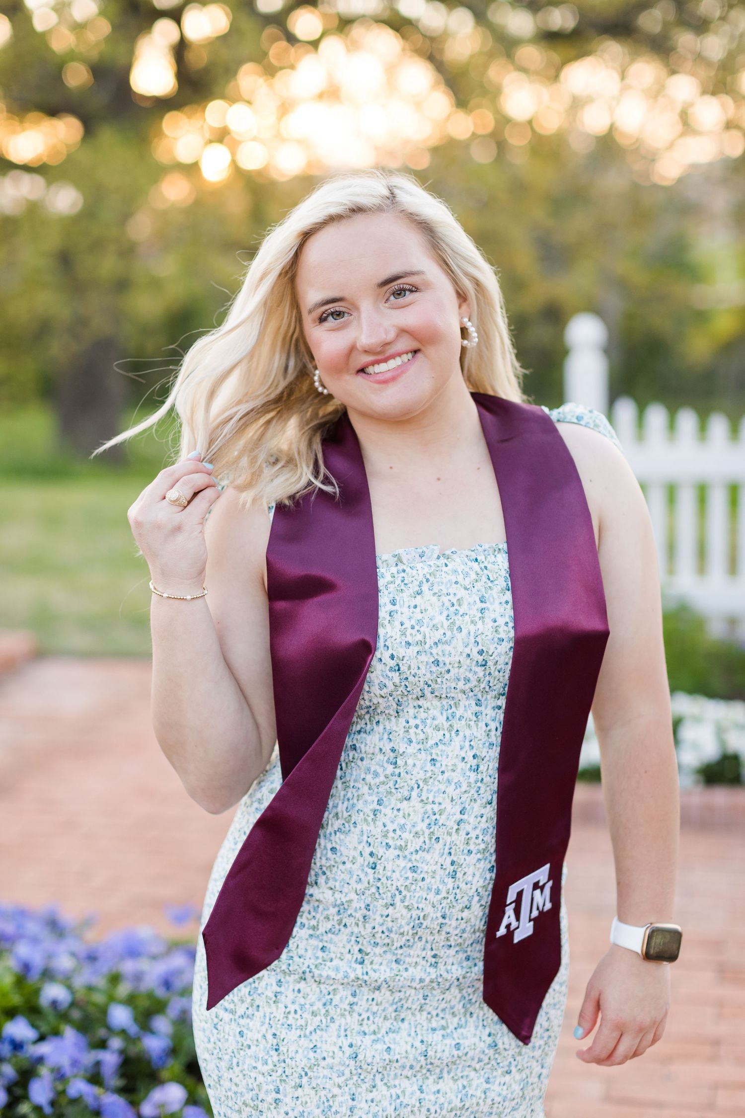 Graduate wearing white floral dress and maroon Texas A&M graduation stole poses in garden with bokeh lighting.