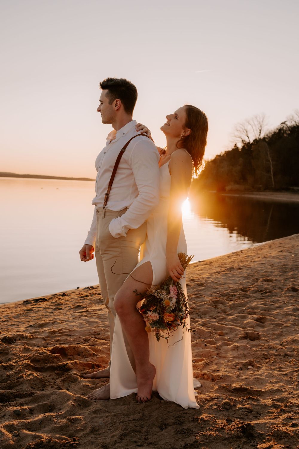 A couple stands back-to-back on a sandy beach at sunset, with the bride holding a bouquet against a serene lake backdrop.