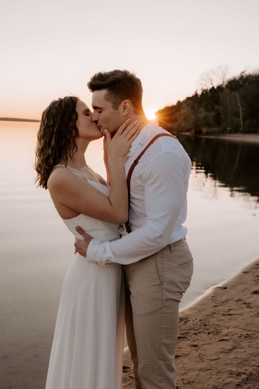 Romantic sunset portrait of a bride and groom sharing an intimate moment on a beach by a lake.