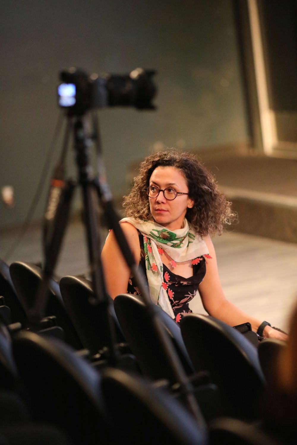 Someone with curly hair and glasses sits in an auditorium with a camera tripod visible in the background.