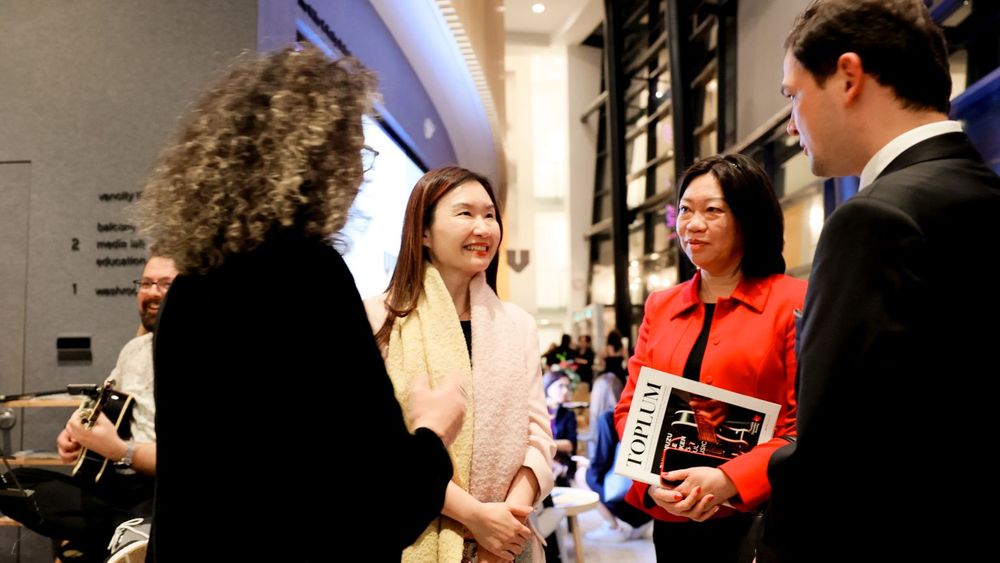 A group engages in lively conversation in an office hallway while holding notebooks and papers.