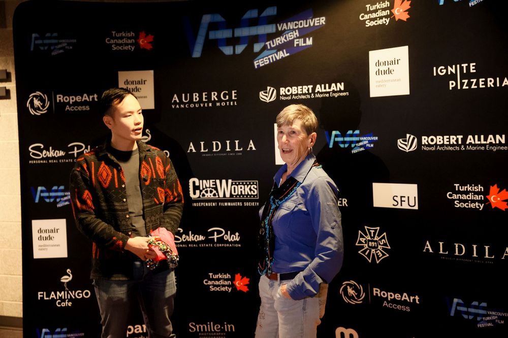 Two people wearing contrasting outfits standing in front of a sponsor wall at a formal event.