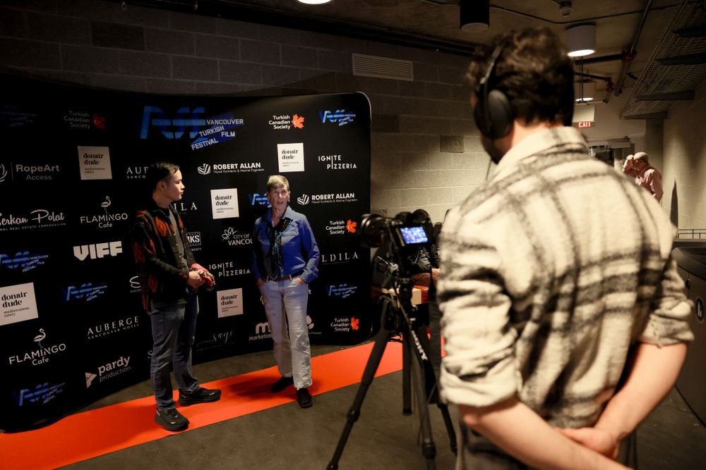 Television interview being filmed on red carpet with camera operator and media backdrop showing sponsor logos.