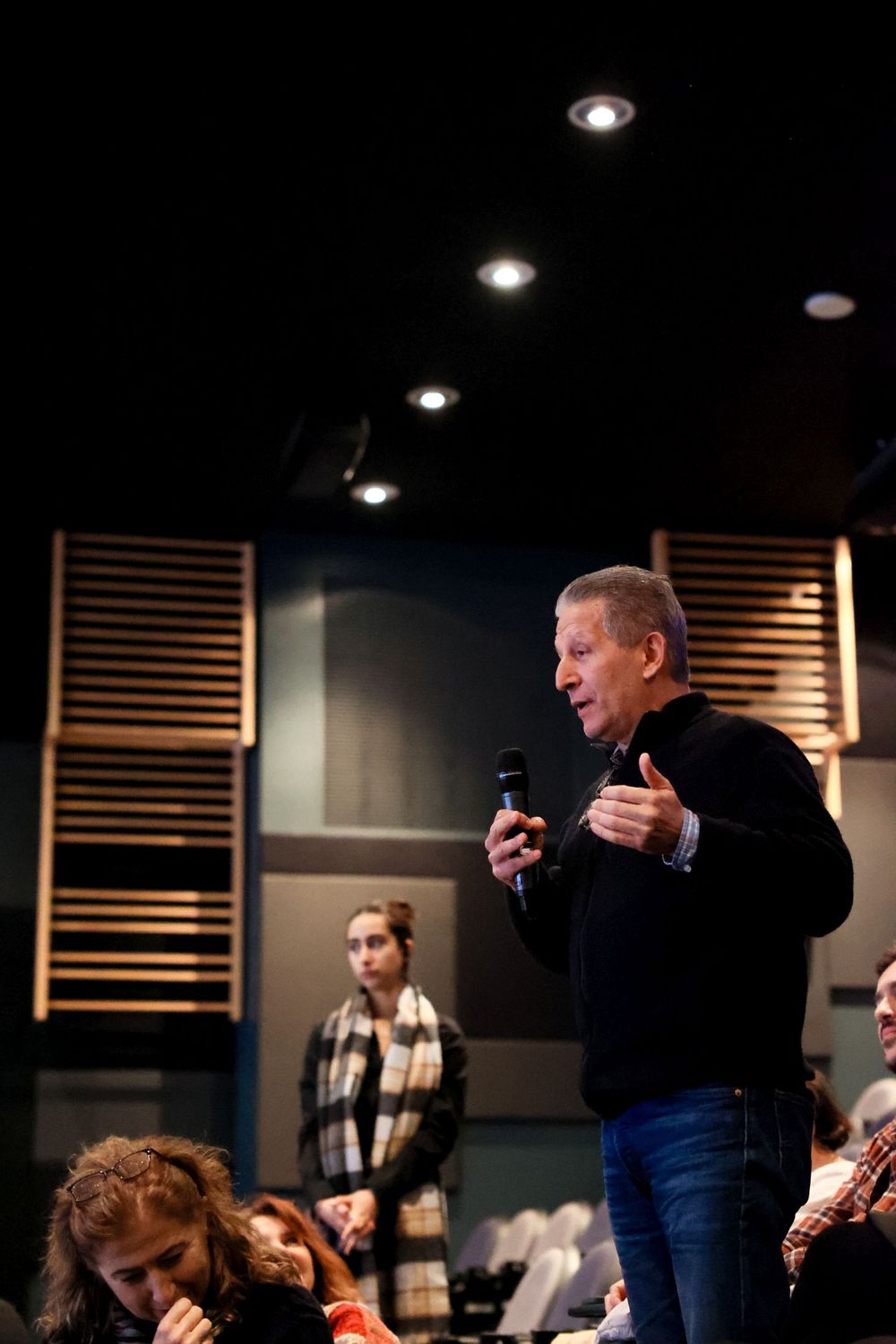 Speaker addressing audience in modern performance space with wooden acoustic panels and spotlights.