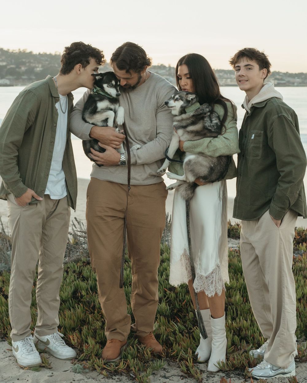 A group of friends in casual attire stand together in a row outdoors against a waterfront backdrop.