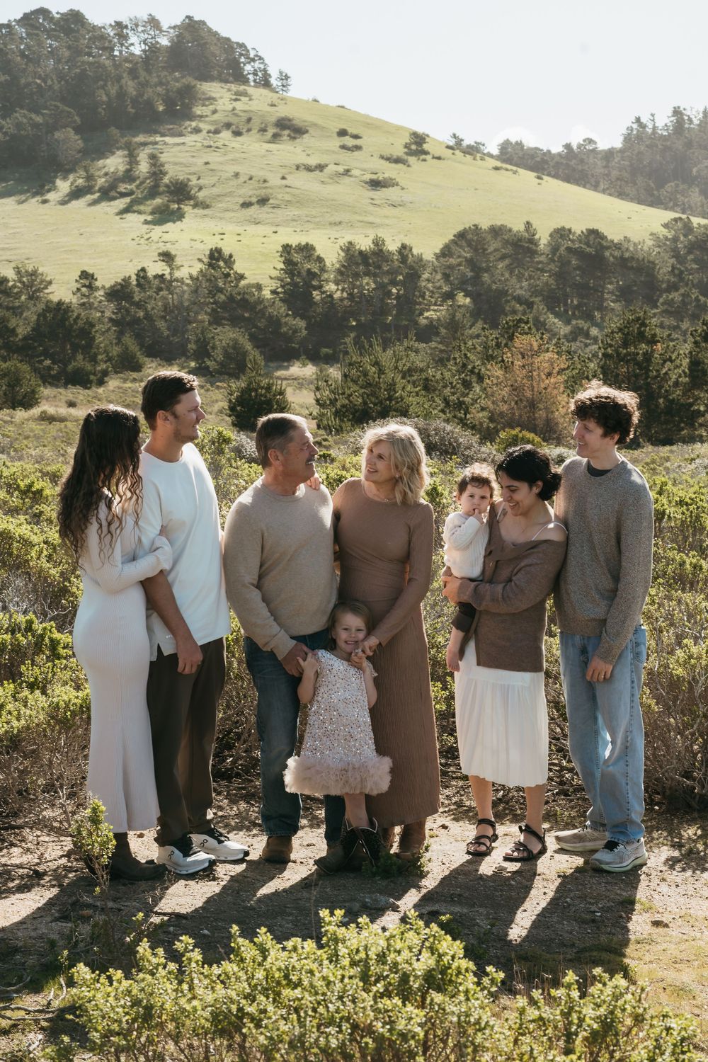 A group of people stand together on a sunny hillside overlooking a scenic natural landscape.