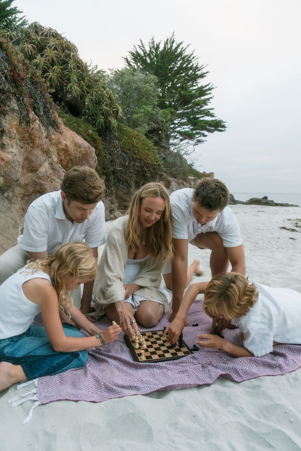 Family playing checkers on a blanket at the beach near rocky cliffs and cypress trees on a cloudy day.