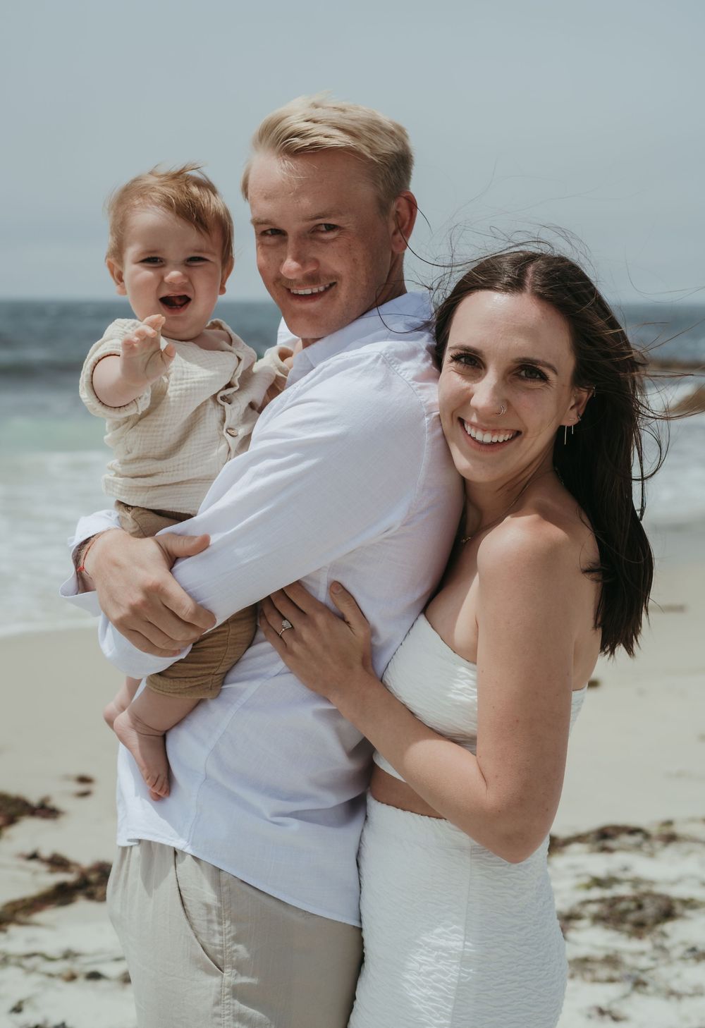 A happy family poses together on a sunny beach with the ocean visible in the background.