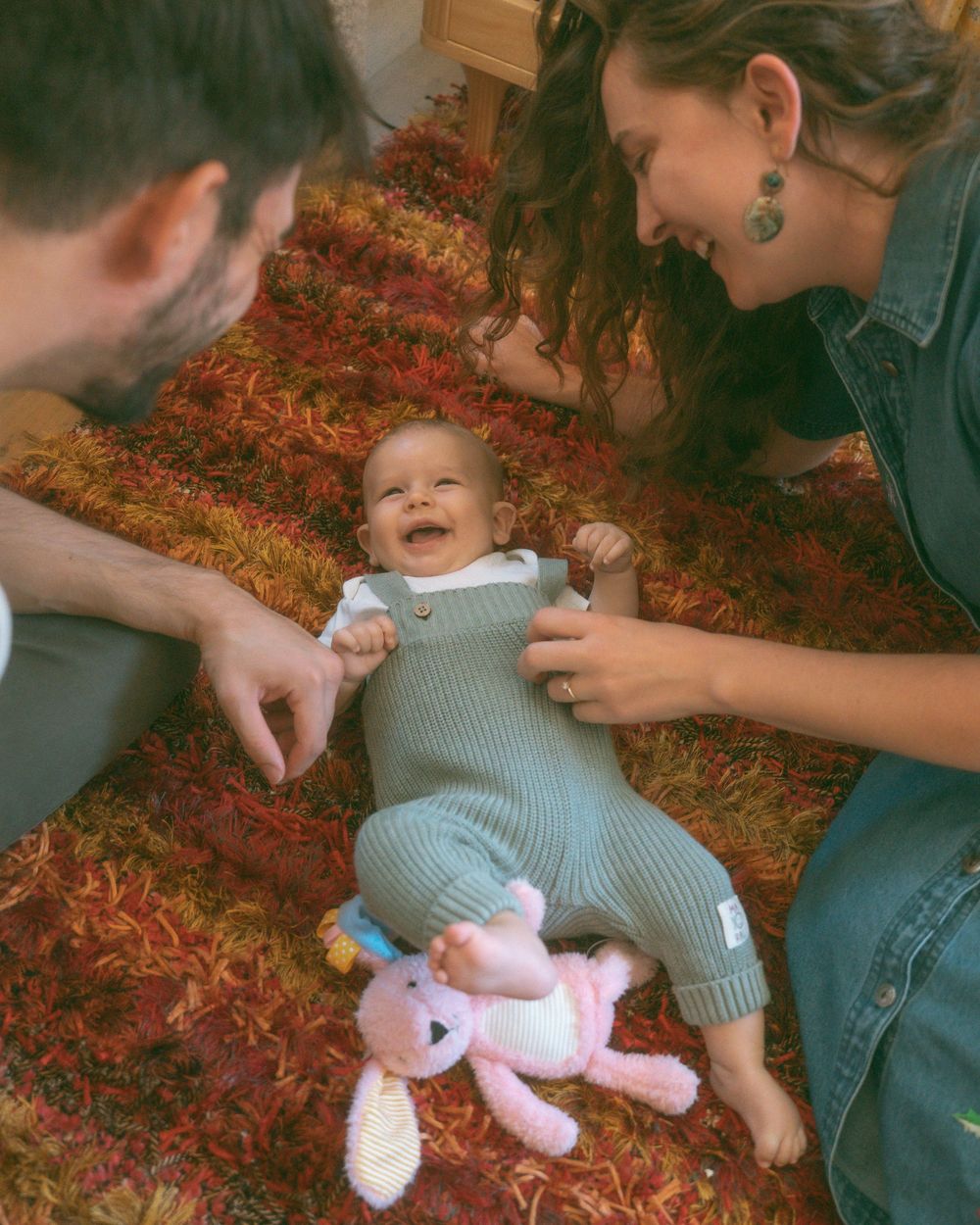 A laughing baby in a green knit romper lies on a colorful rug between two smiling parents with a pink unicorn toy.