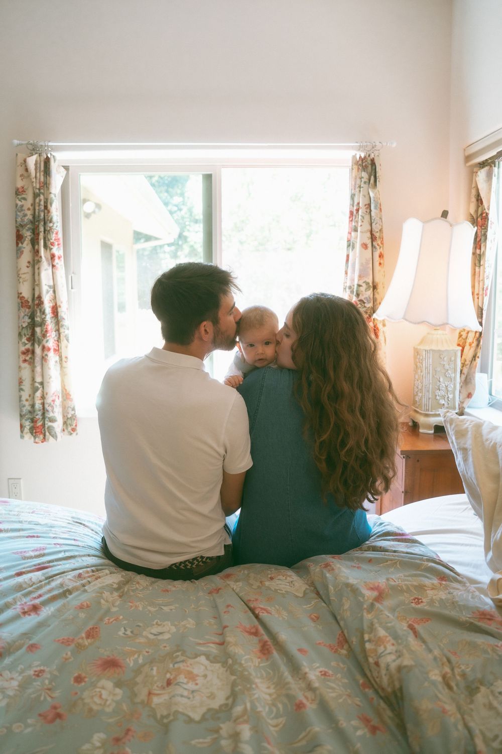 A couple sits on a floral bed kissing their baby, viewed from behind near a bright window with curtains.