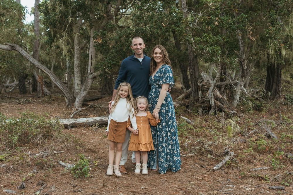 A family poses together in a wooded outdoor setting wearing coordinated blue and brown outfits.
