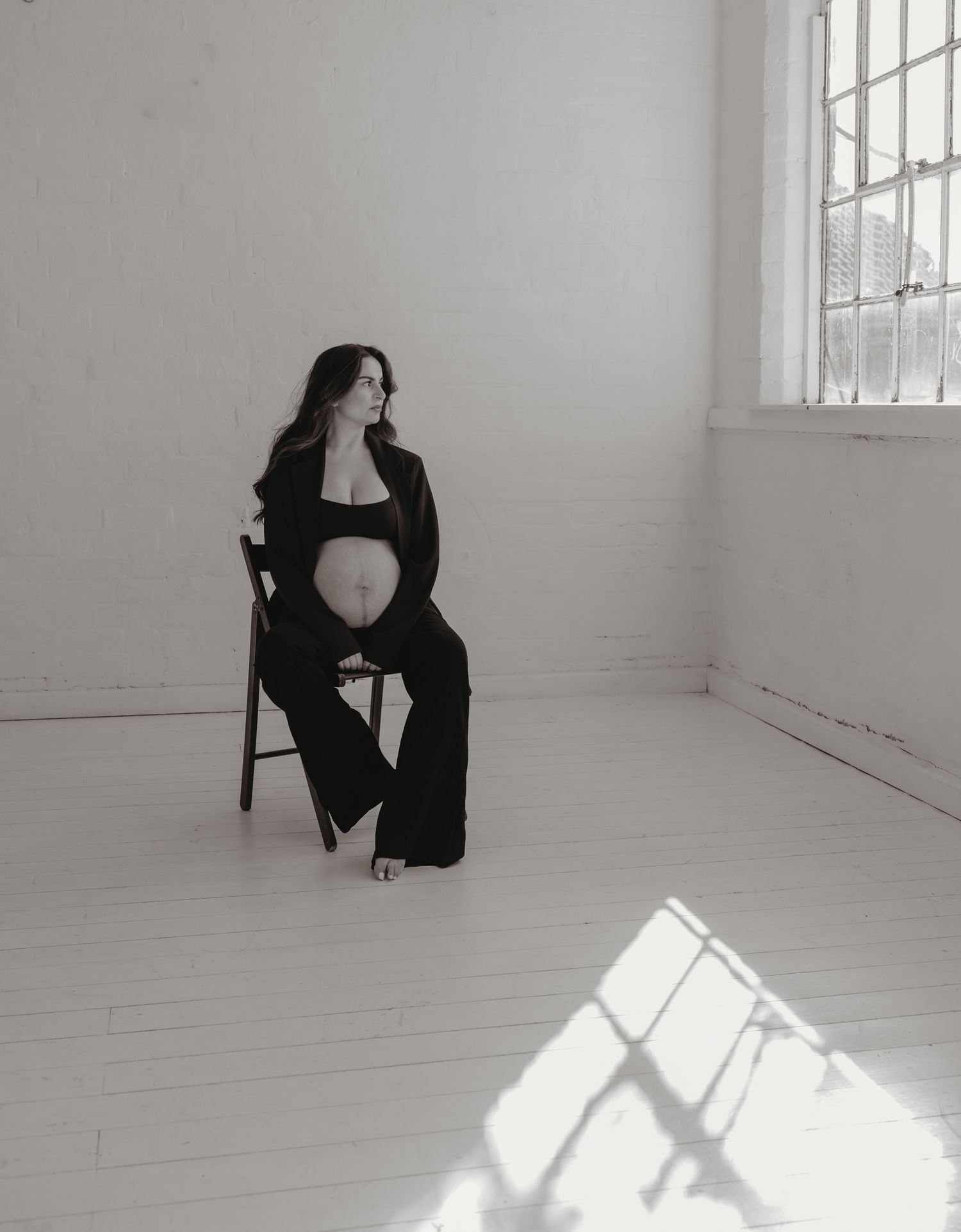 Leicestershire maternity photographer. Portrait of an expectant mother seated in a white room with dramatic window lighting casting shadows.