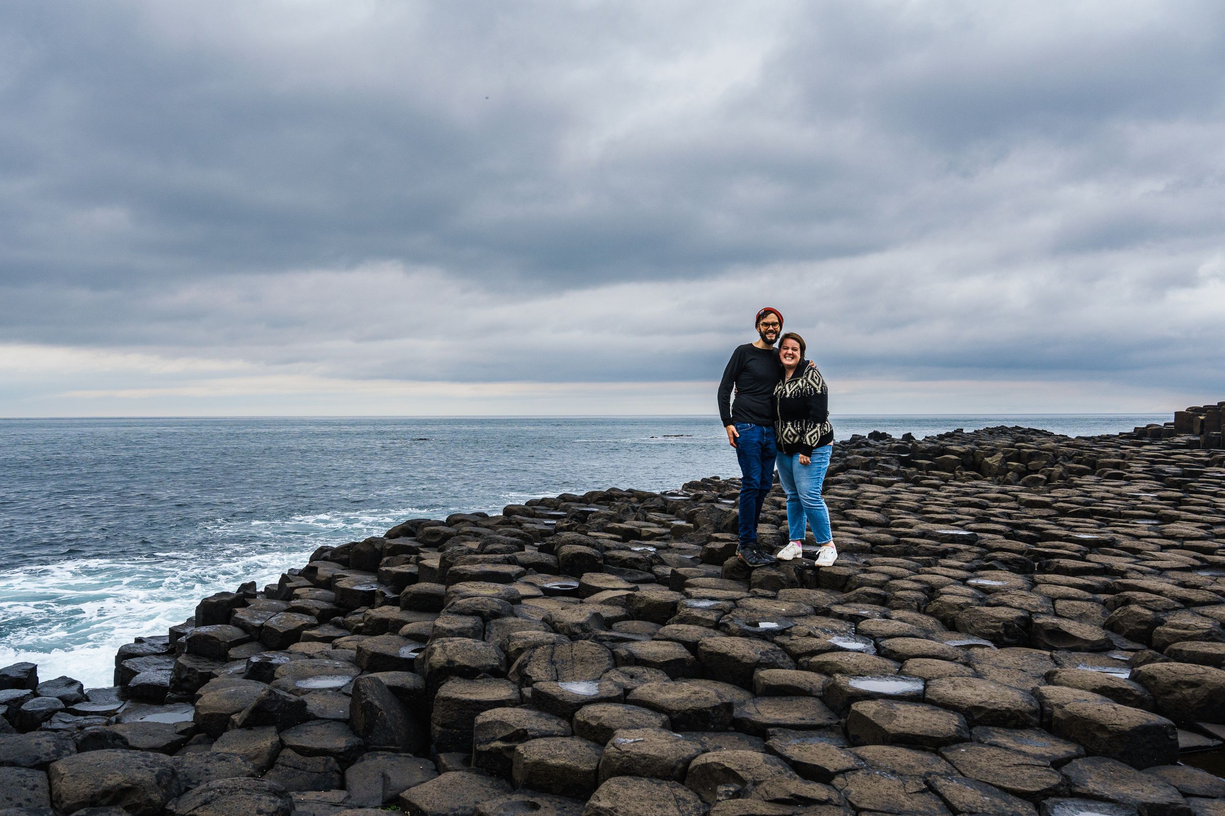 Two people stand on the dramatic hexagonal basalt columns of Giant's Causeway in Northern Ireland under moody skies.