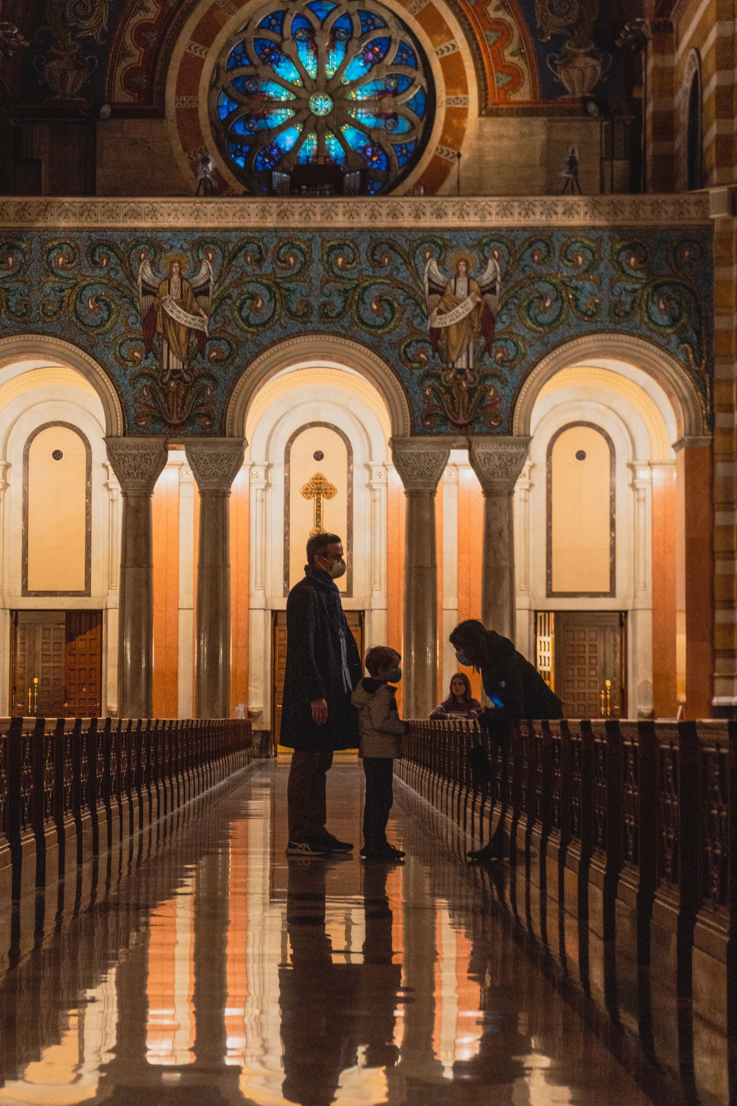 Two silhouettes stand in ornate cathedral aisle with stained glass rose window and arched architecture reflecting on marble floor.