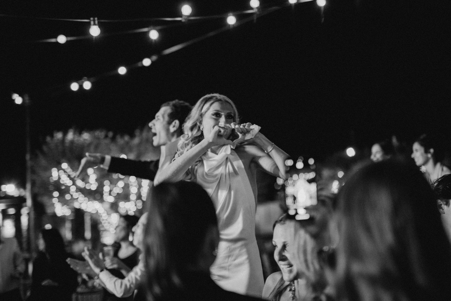 Black and white photo of concert crowd enjoying live music performance at nighttime with string lights overhead.