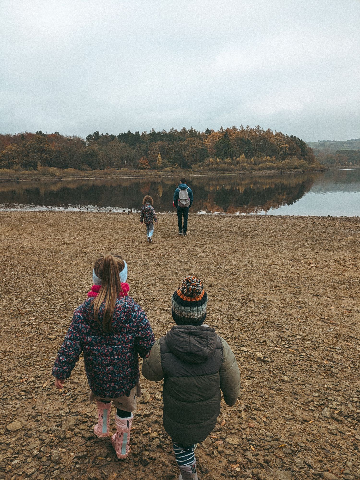 People walking along a rocky shoreline at a tranquil lake surrounded by autumn trees on a cloudy day.