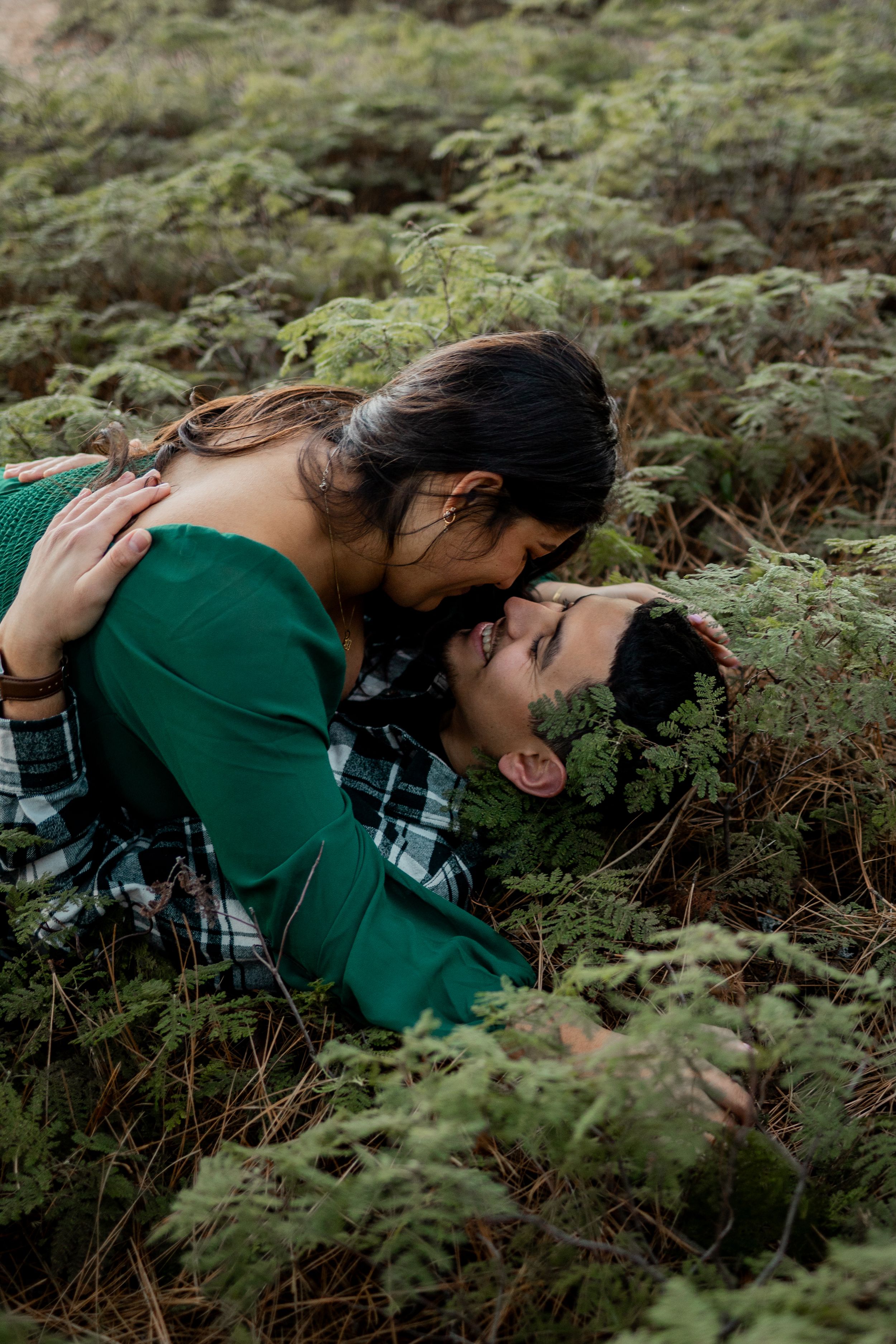 A couple shares a romantic moment lying in lush green ferns, with the woman leaning down to kiss her partner outdoors.