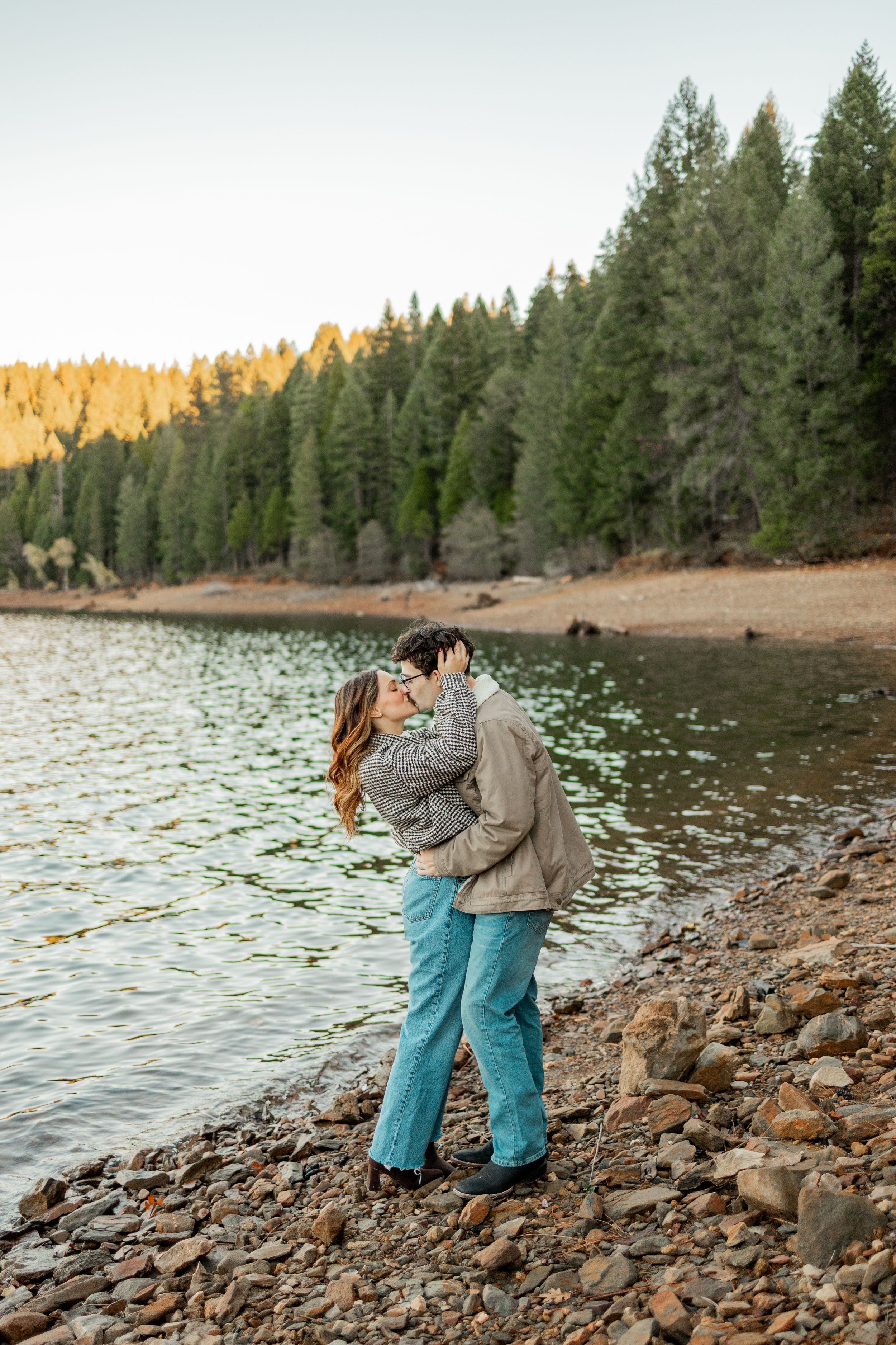 Couple kissing on rocky lakeside shore surrounded by tall pine trees at golden hour.