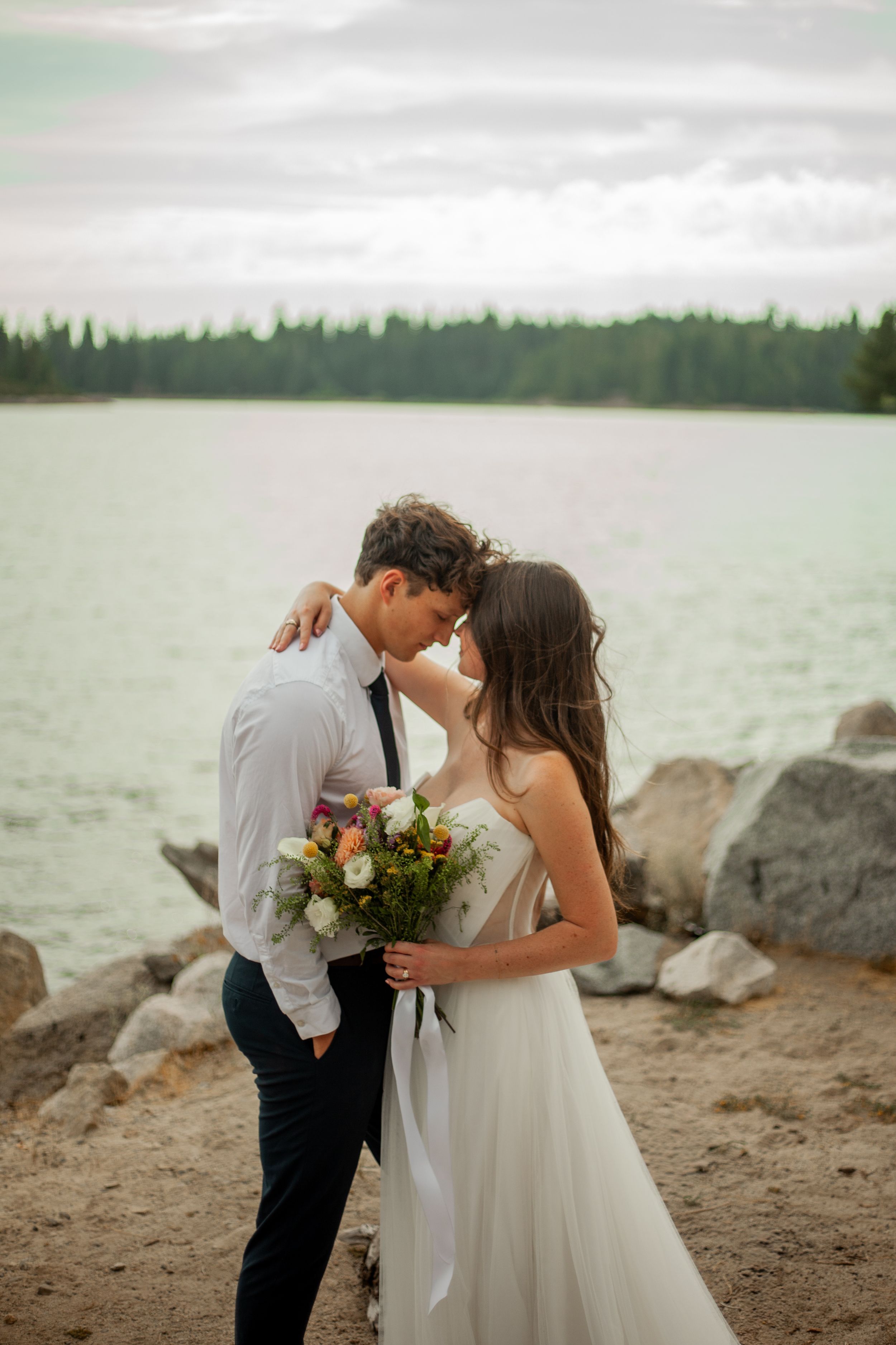 Bride and groom share intimate moment by lakeside, she holds colorful bouquet in white gown, he wears white shirt and black tie.