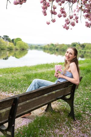 À Huy, portrait de femme photographiée en extérieur, assise sur un banc au bord de l’eau avec un cerisier japonais en fleur au printemps