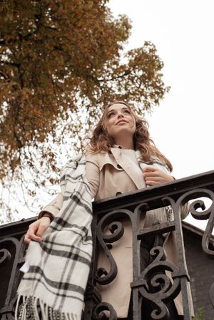 À Liège, portrait de femme photographiée en extérieur en automne près d’un arbre et d’une barrière, tenant son écharpe avec assurance