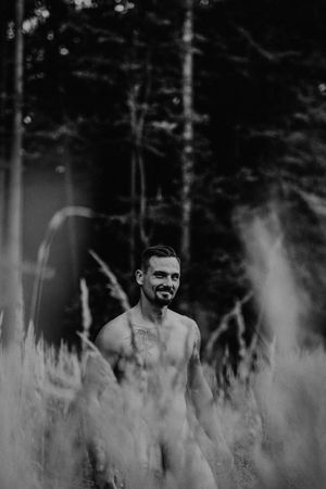 Black and white portrait of a person smiling while standing in tall grass with trees in the background.