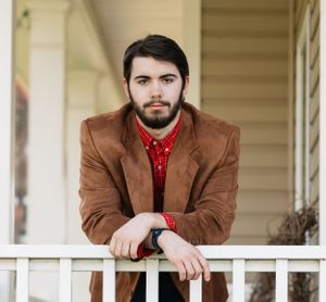 Portrait on white porch railing wearing brown blazer and red button-down shirt.
