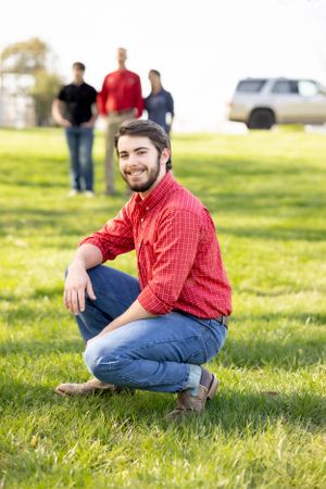 Person in red plaid shirt and jeans kneels on grassy field with others visible in background.