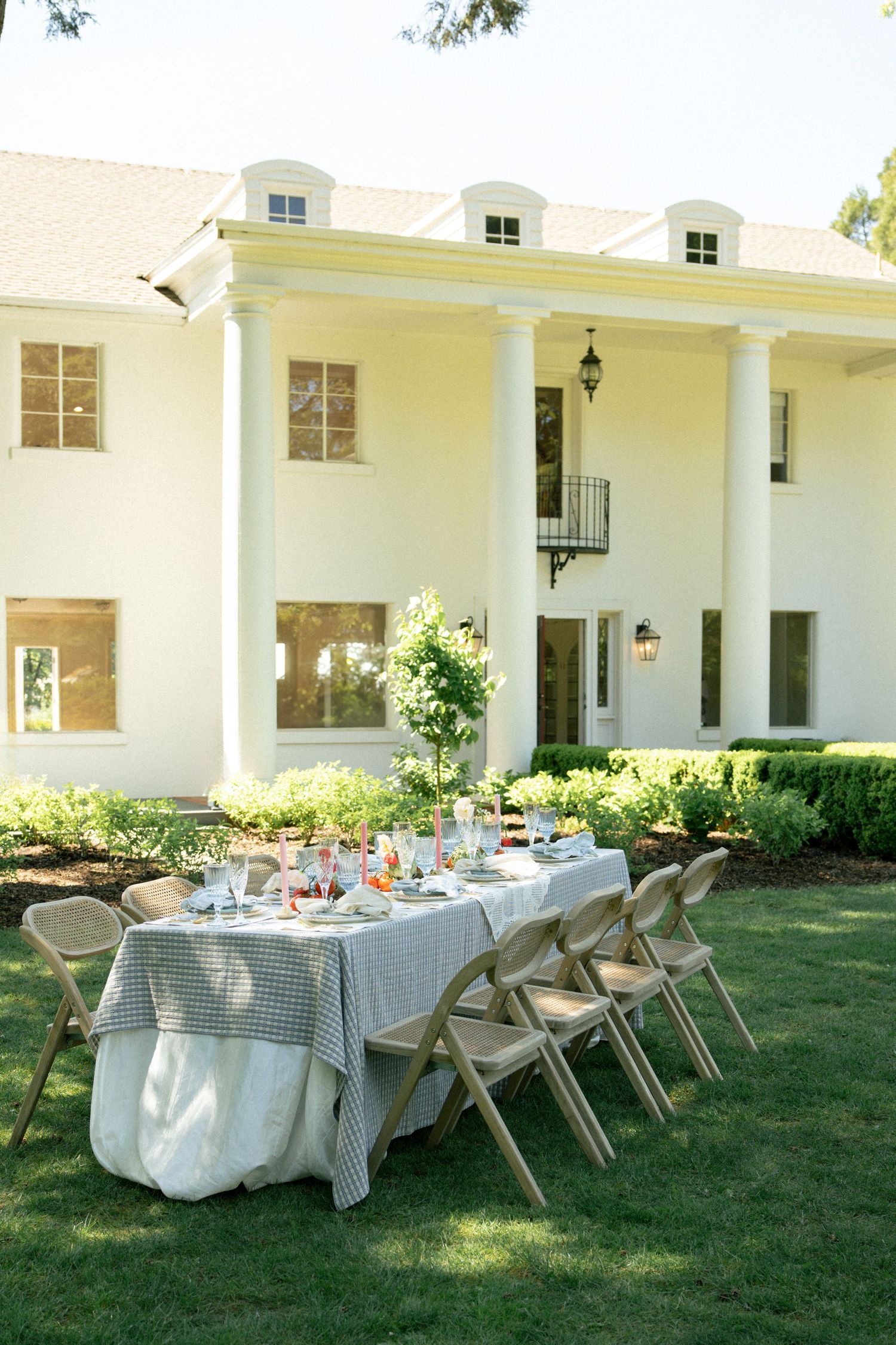 Elegant outdoor dining table set with gingham tablecloth on green lawn before a grand white colonial mansion.