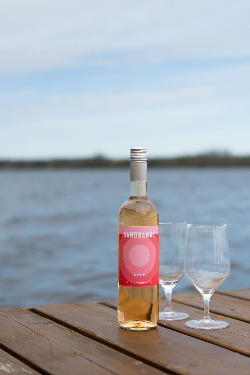 Bottle of rose wine with two glasses on wooden dock overlooking water.